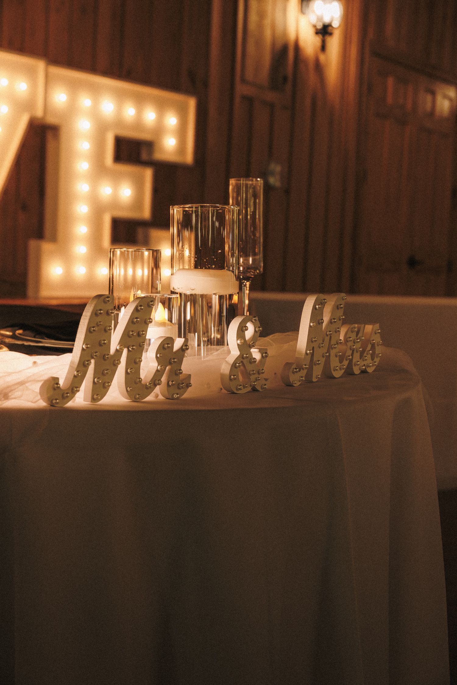 Wedding sweetheart table with Mr. & Mrs. signs, candles, and illuminated marquee letters in warm romantic lighting.