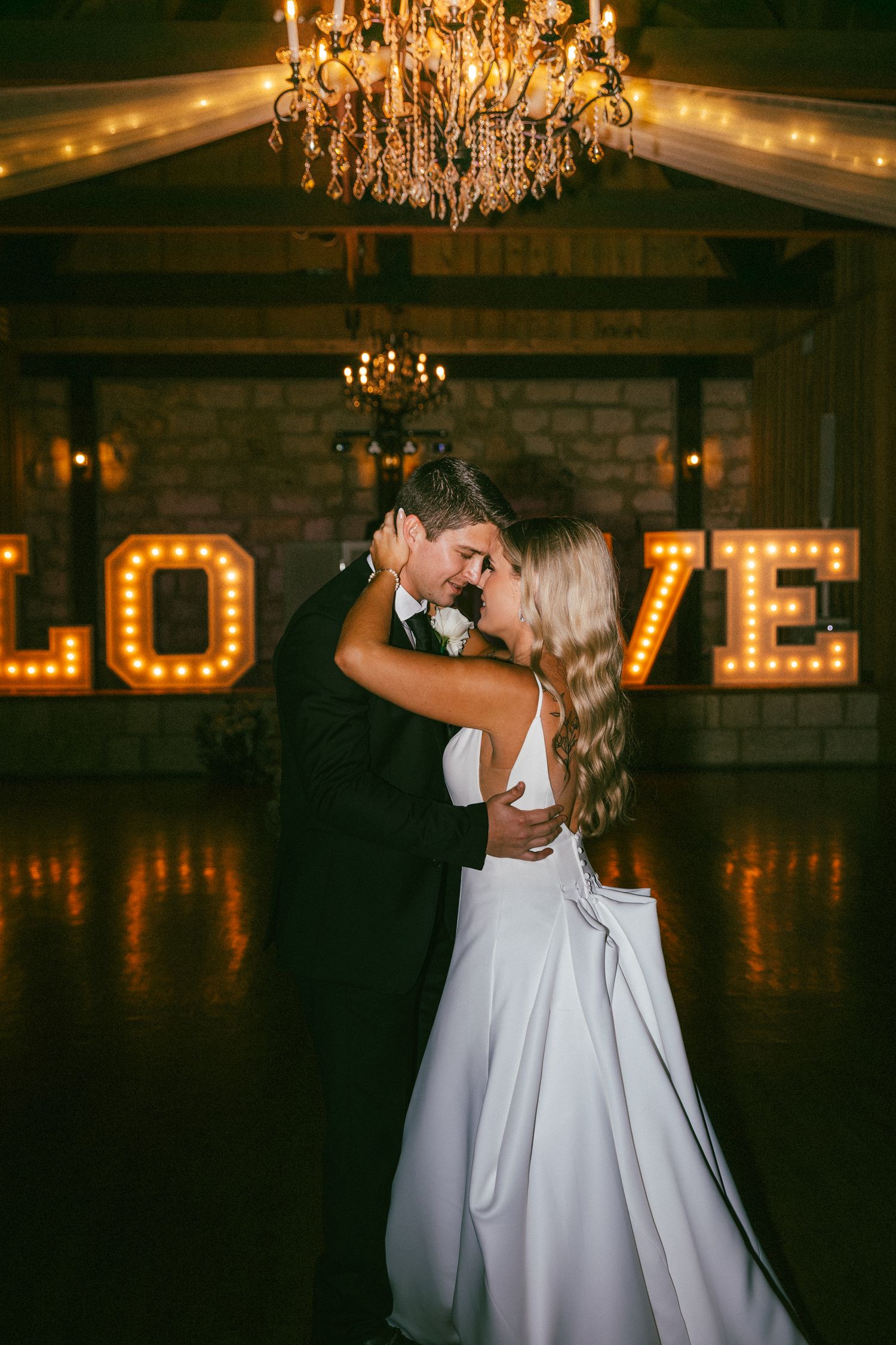 Bride and groom share first dance beneath a chandelier with illuminated LOVE marquee letters.