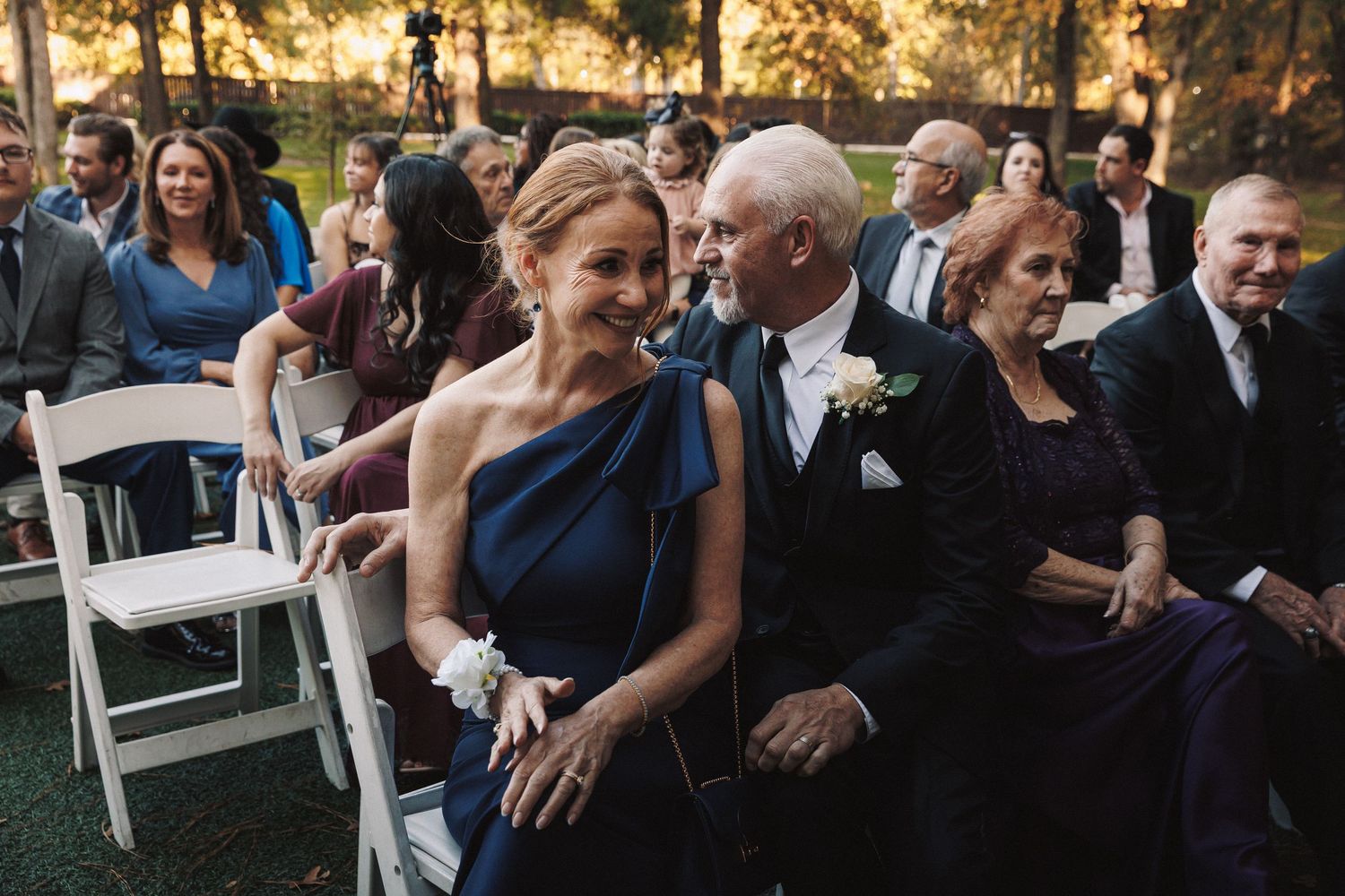 Smiling couple in formal attire seated at an outdoor wedding ceremony among guests on white chairs.
