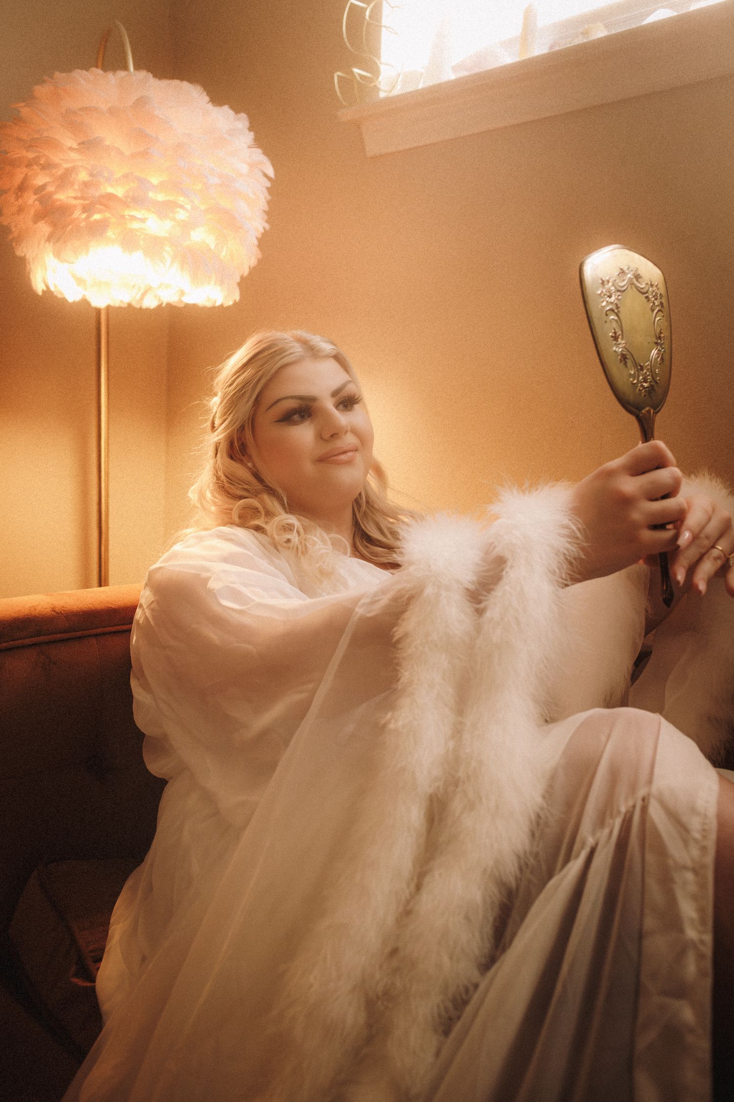 Blonde bride in white fur robe holding ornate hand mirror on velvet sofa with feather lamp.