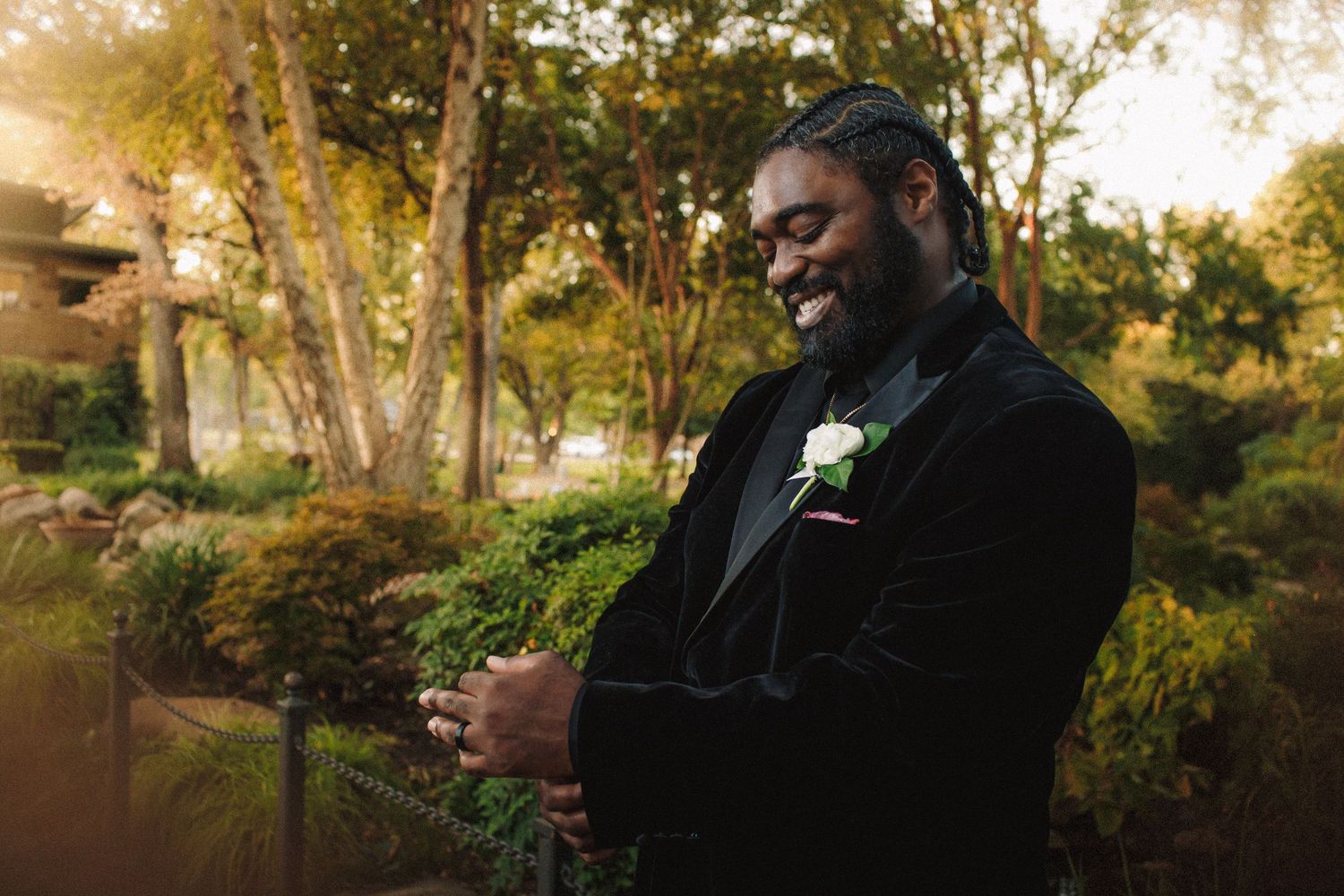 Smiling groom in black velvet tuxedo with boutonniere standing in lush garden at golden hour.