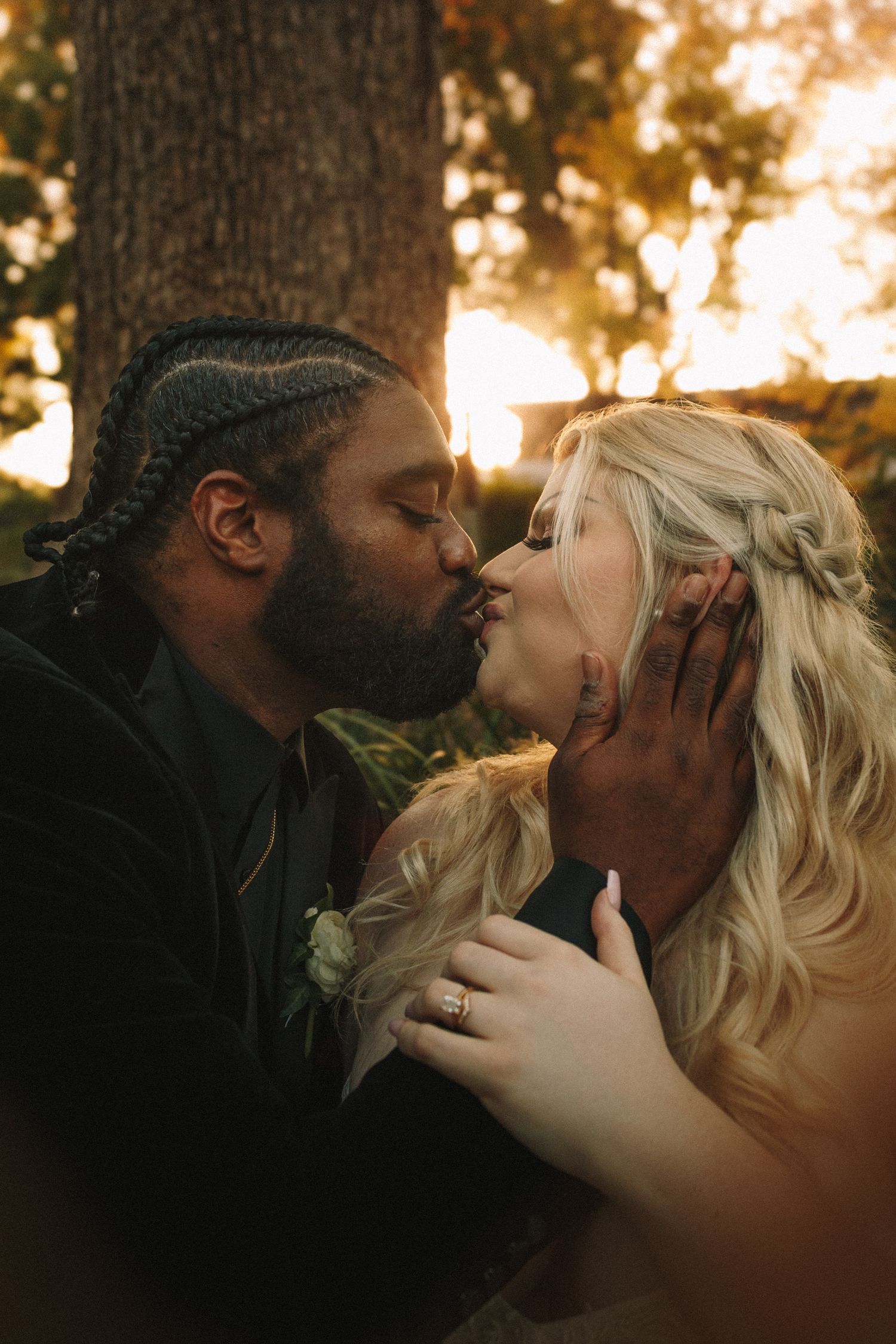 Groom with braided hair kisses bride in lace gown beneath a sunlit tree at golden hour.