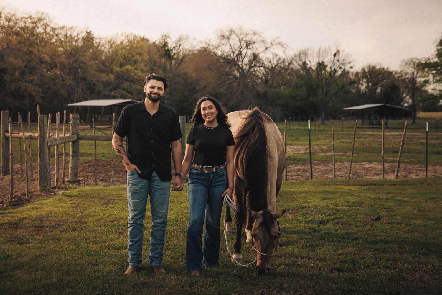 Couple standing hand in hand beside a horse on a farm at golden hour.