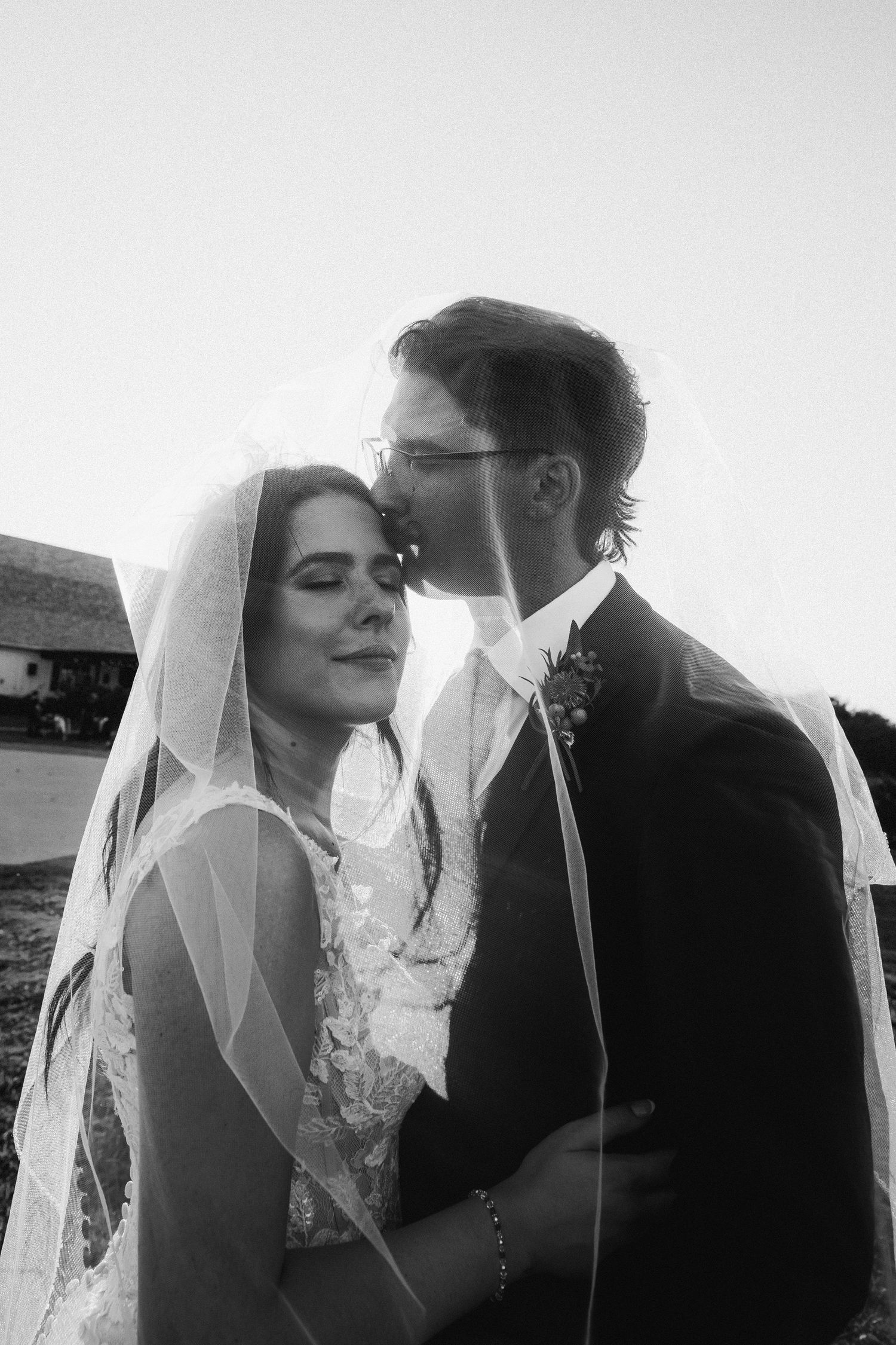 Black and white wedding photo of bride and groom sharing tender kiss under flowing veil.