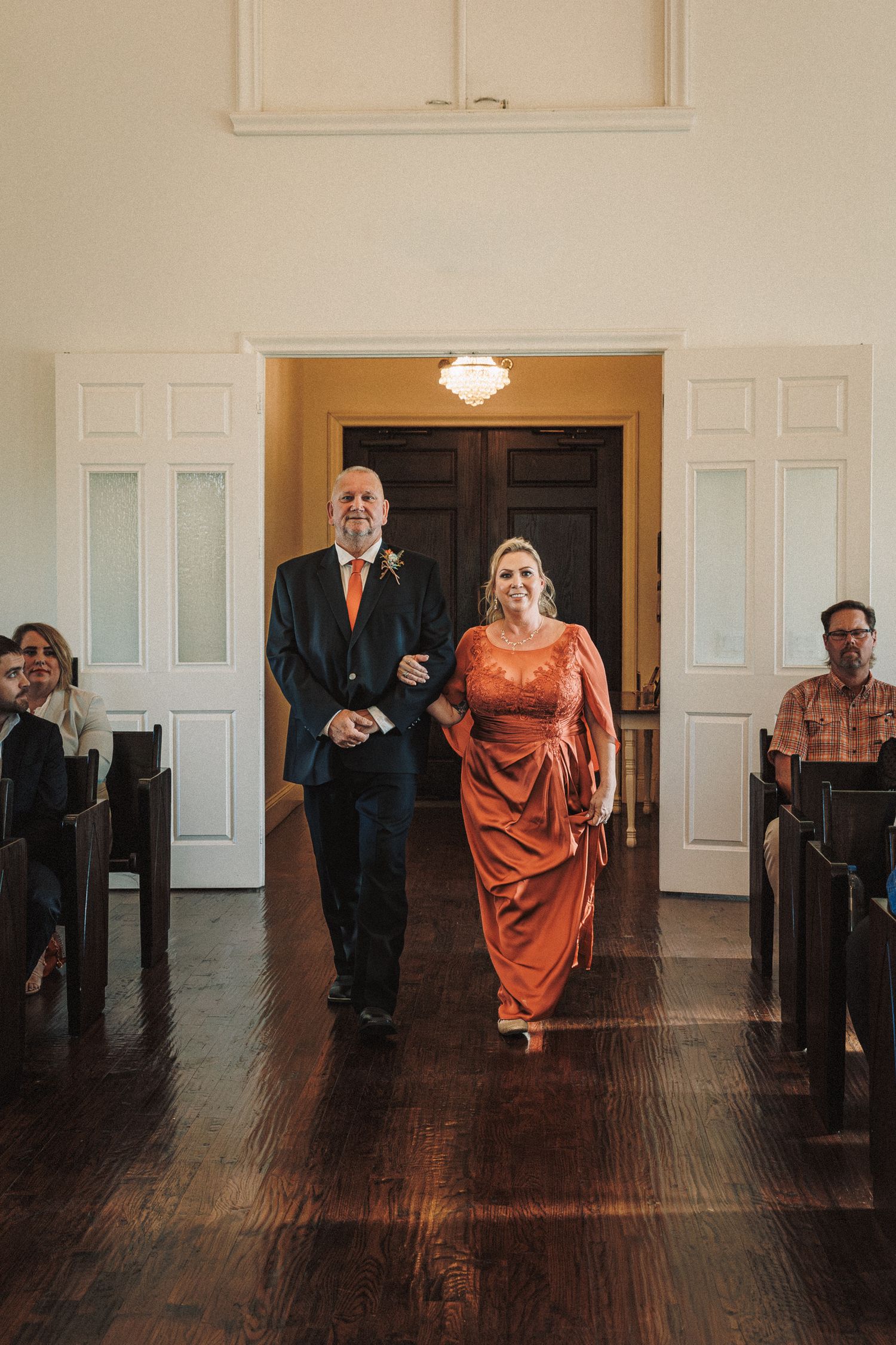 Couple walking down church aisle, man in navy suit and woman in elegant orange gown at wedding.