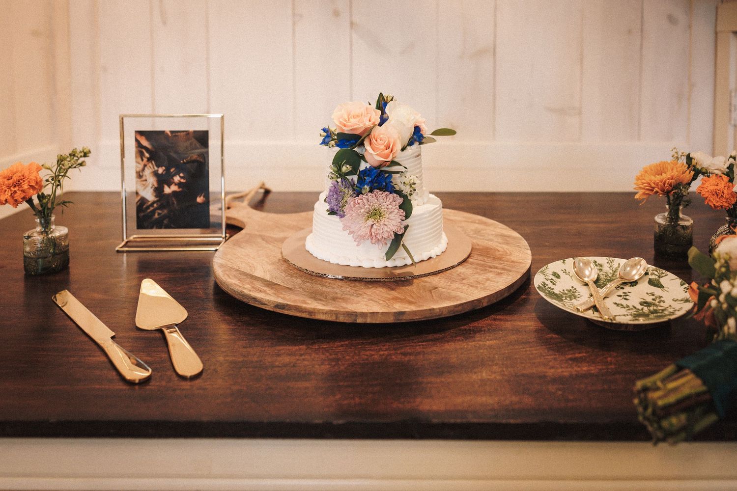 Elegant wedding cake with floral topping on wooden board with cake serving set and orange flowers.