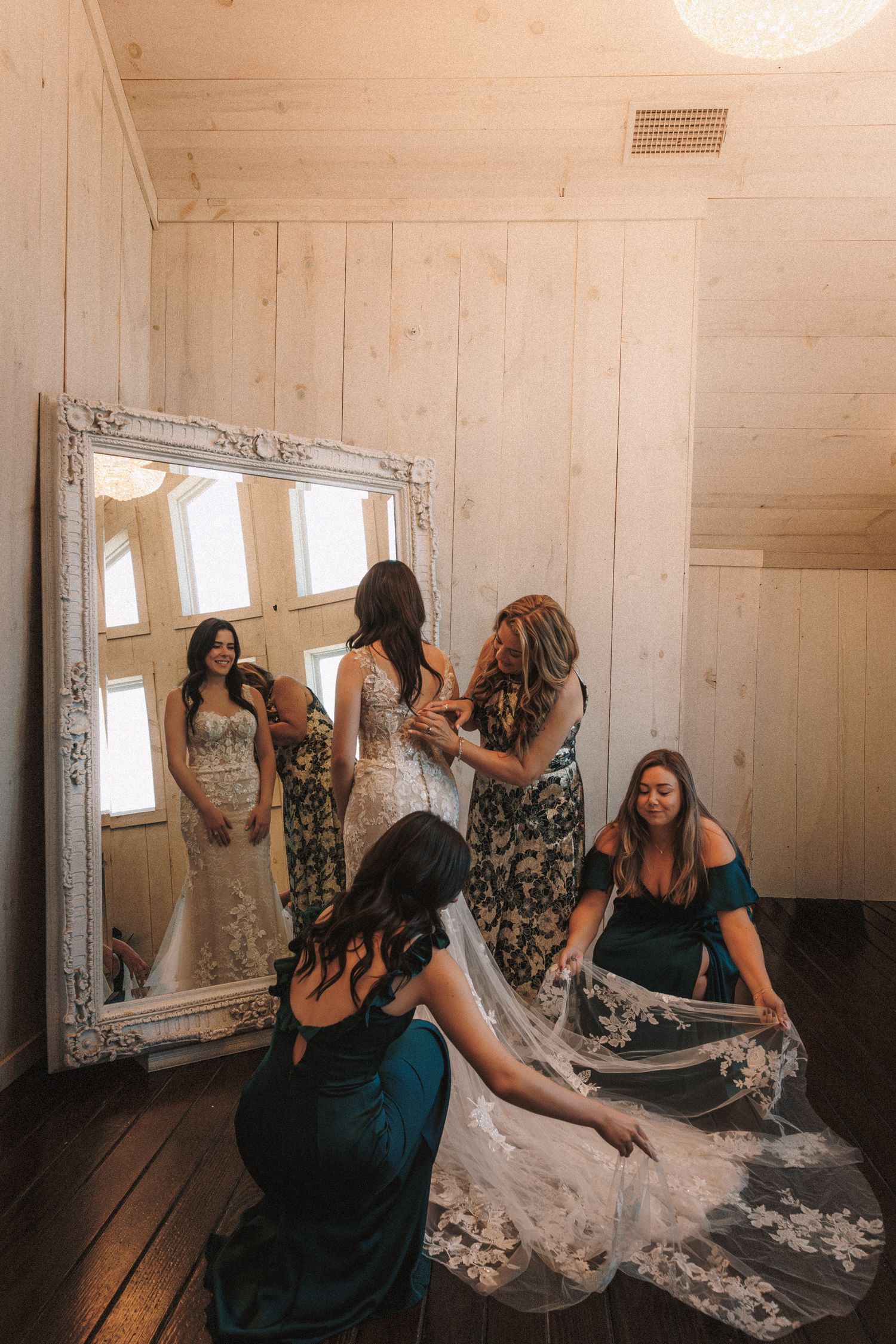 Bridesmaids in teal dresses helping a bride with her lace veil before a large ornate mirror.