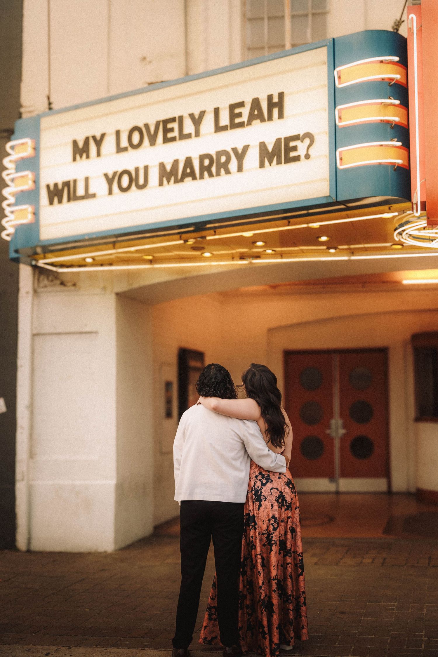 Couple embraces under lit theater marquee reading My Lovely Leah Will You Marry Me at night.