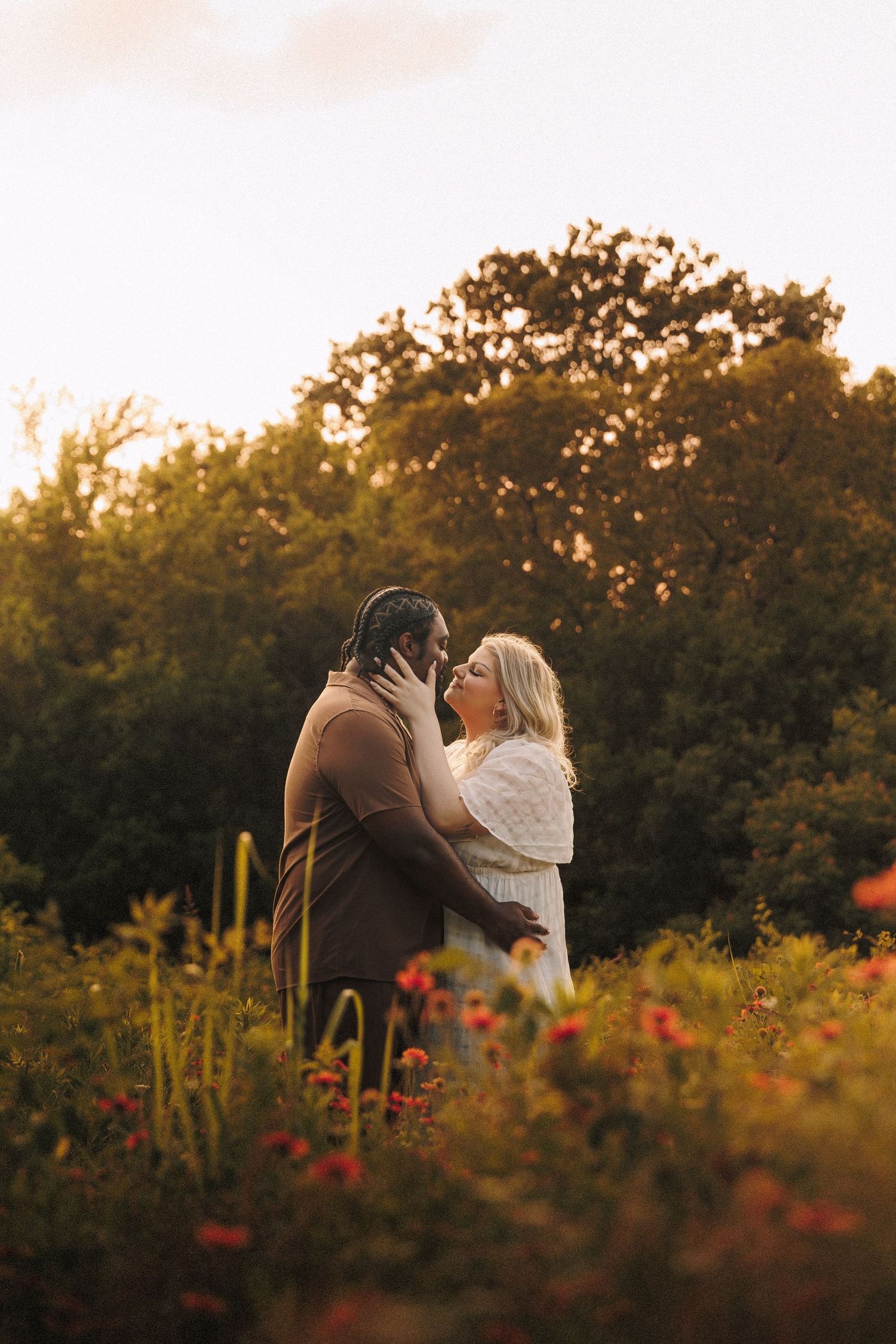 A couple embraces in a romantic sunset garden with red flowers in the foreground and golden light streaming through trees.