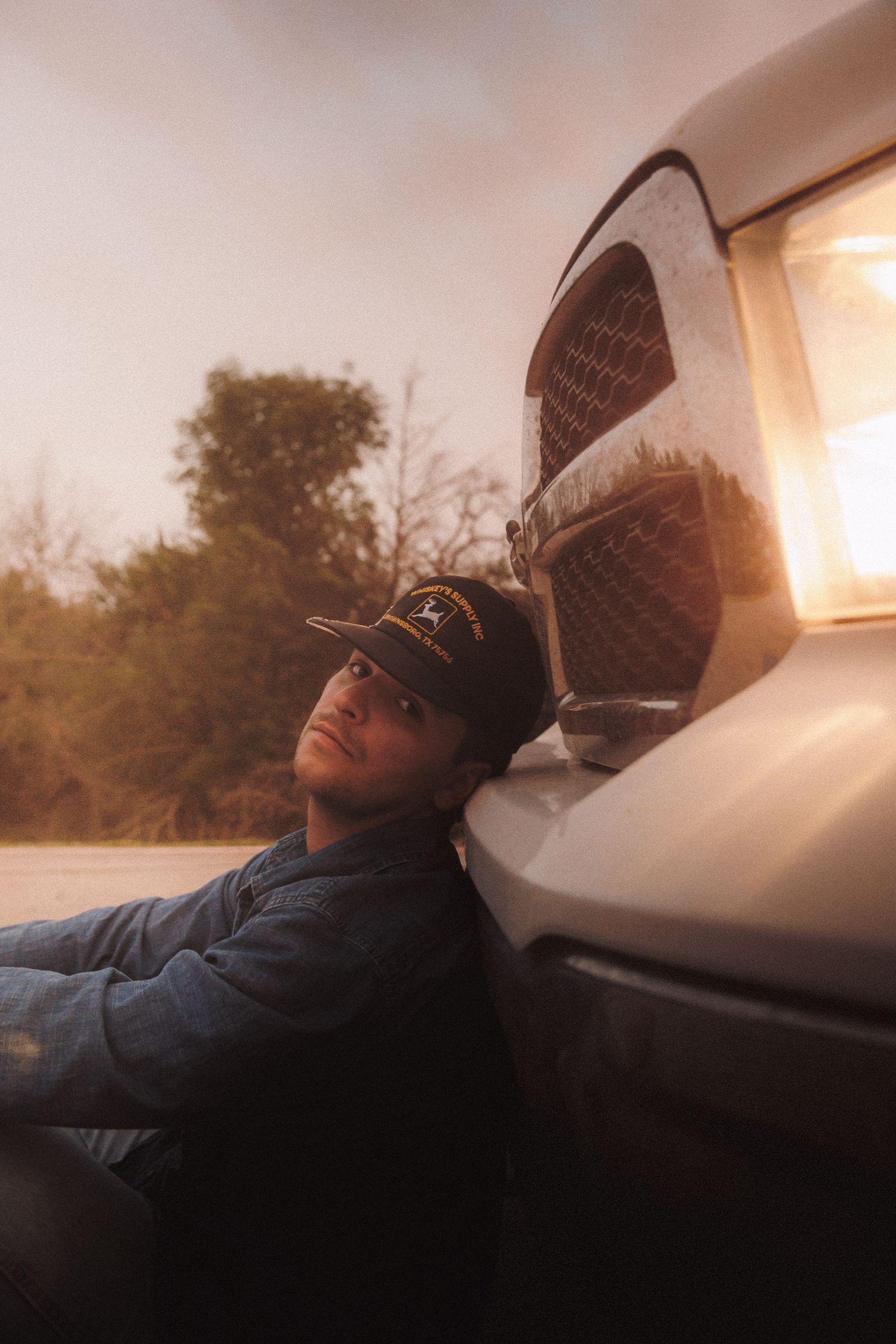Person wearing winter hat relaxes against car window at sunset with warm golden light streaming in.