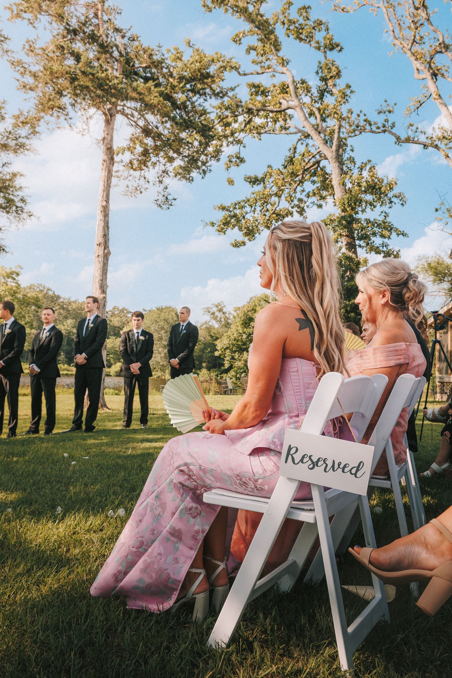 White wooden chairs set up for an outdoor wedding with guests seated under tall trees.