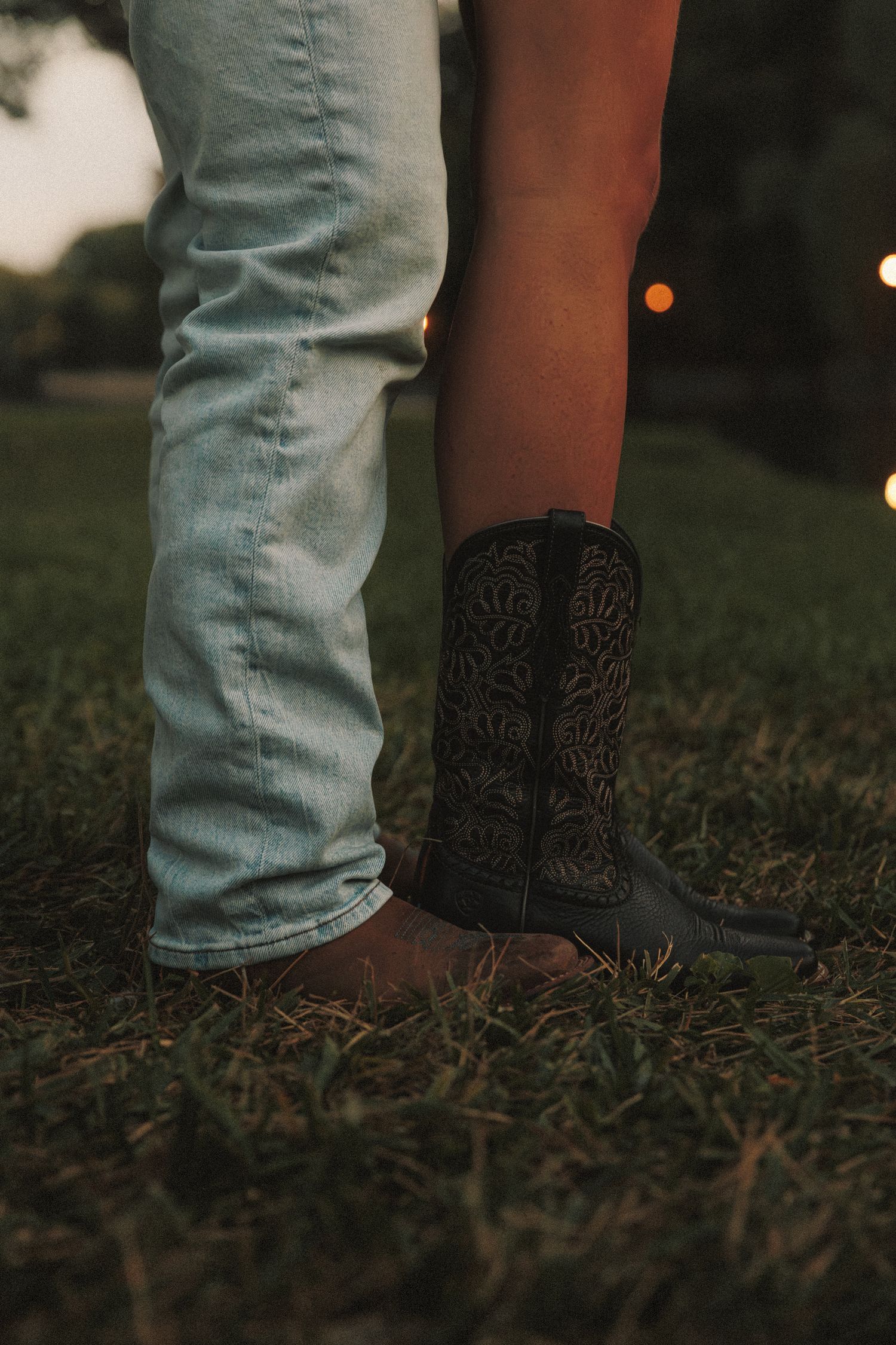 Light denim jeans and black cowboy boots stand together on grass at dusk.
