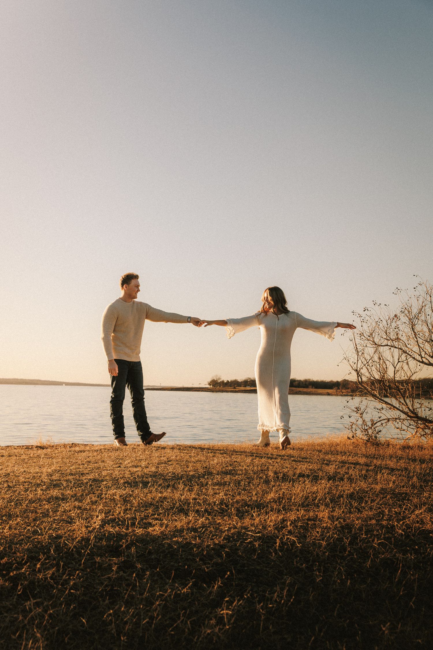 A couple stands silhouetted against a golden sunset on a grassy hilltop overlooking a serene waterfront.