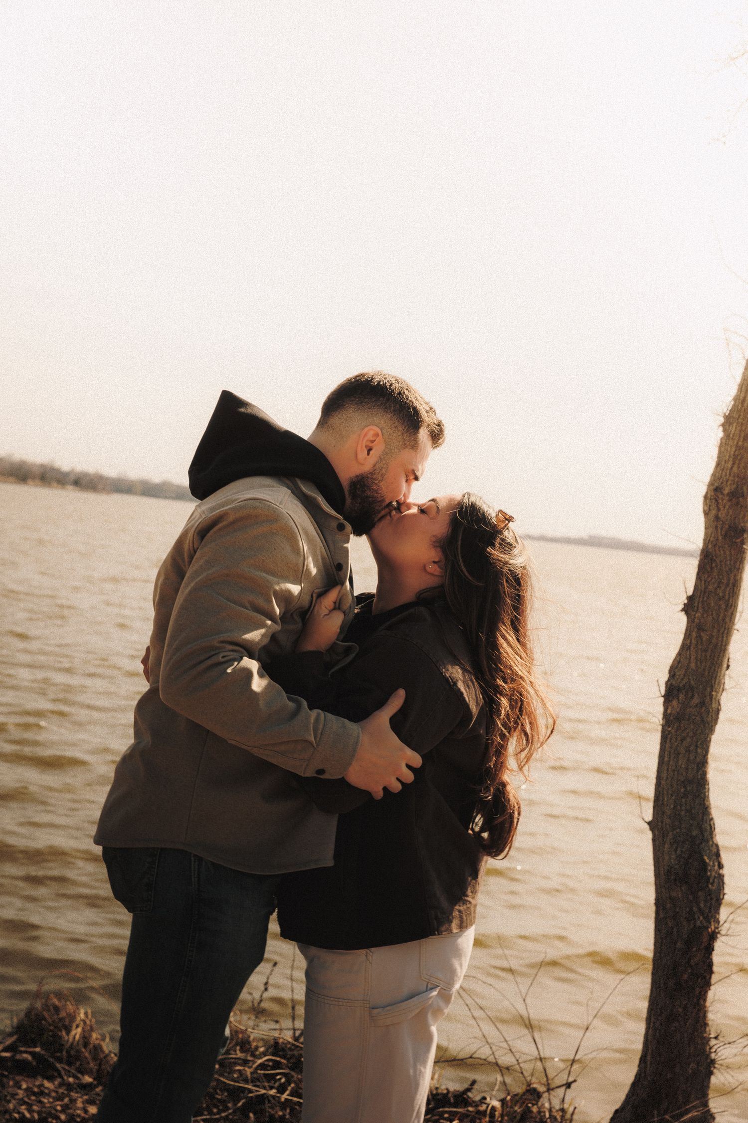 Romantic couple sharing an intimate embrace by the waterfront at sunset, wearing casual winter outerwear.