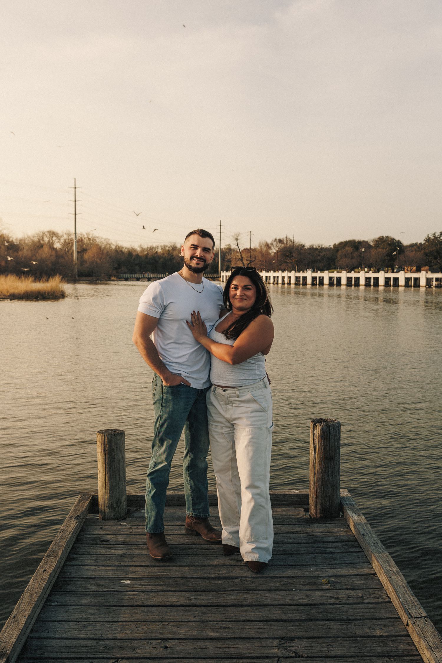 A couple poses together on a wooden pier with water and boats in background.