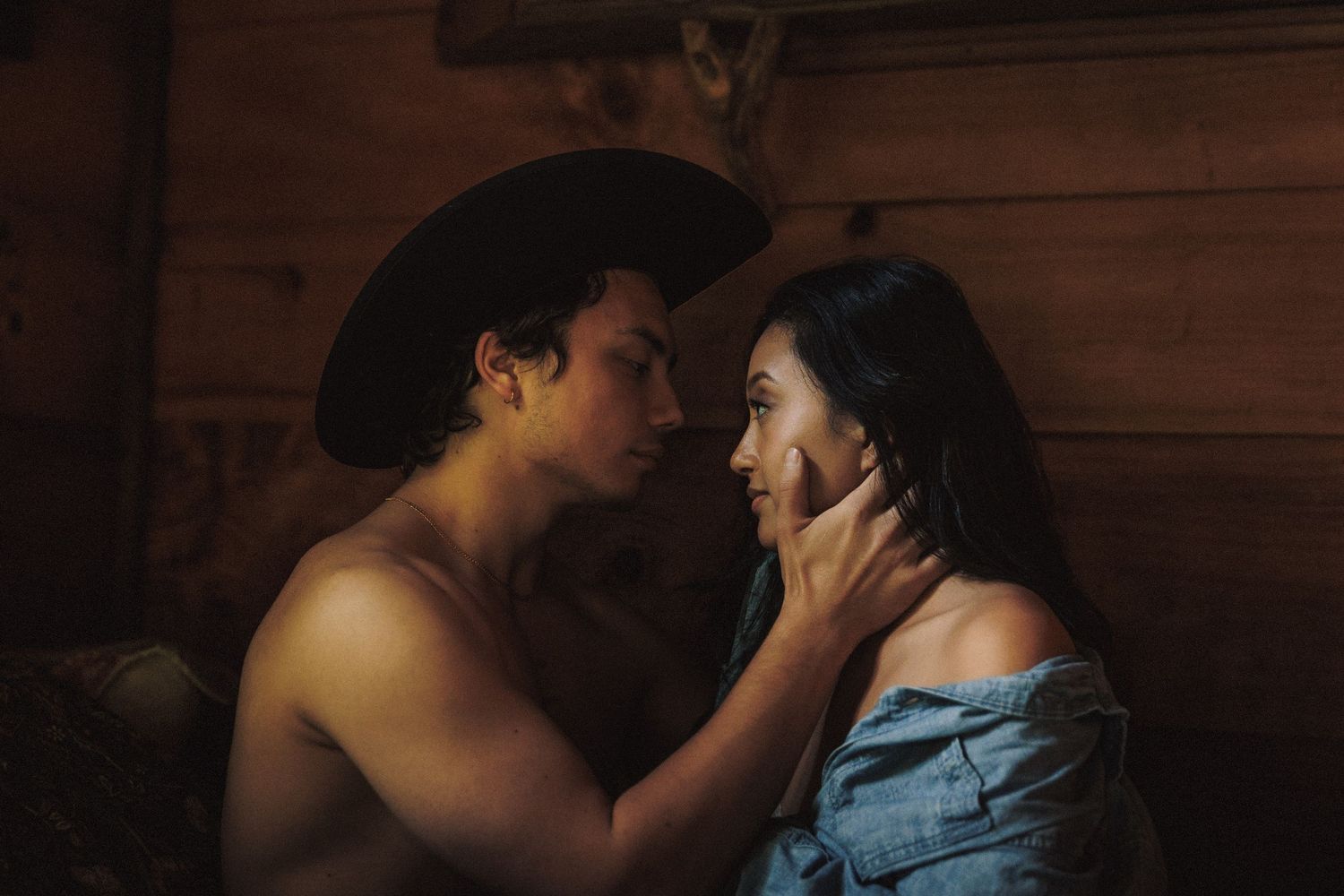 A romantic couple share an intimate moment in dim lighting against a wooden wall backdrop.