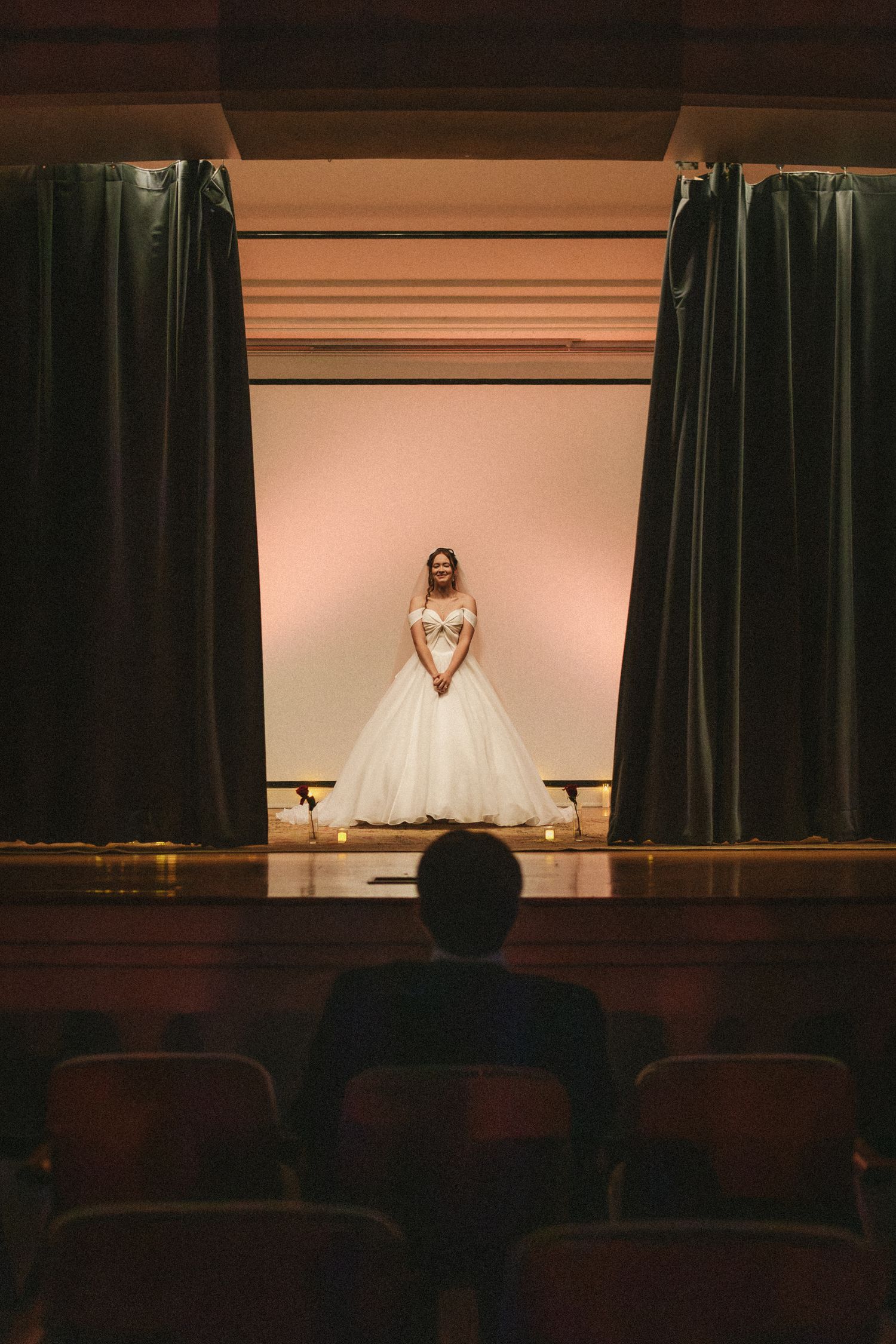 Bride in white off-shoulder ballgown stands on stage as groom watches from theater seats.