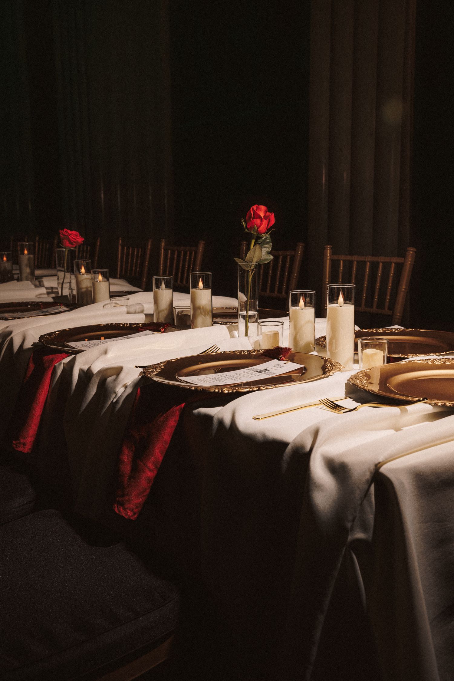 Elegant wedding table setting with white linens, red roses, gold chargers, and glowing candles in a dimly lit venue.