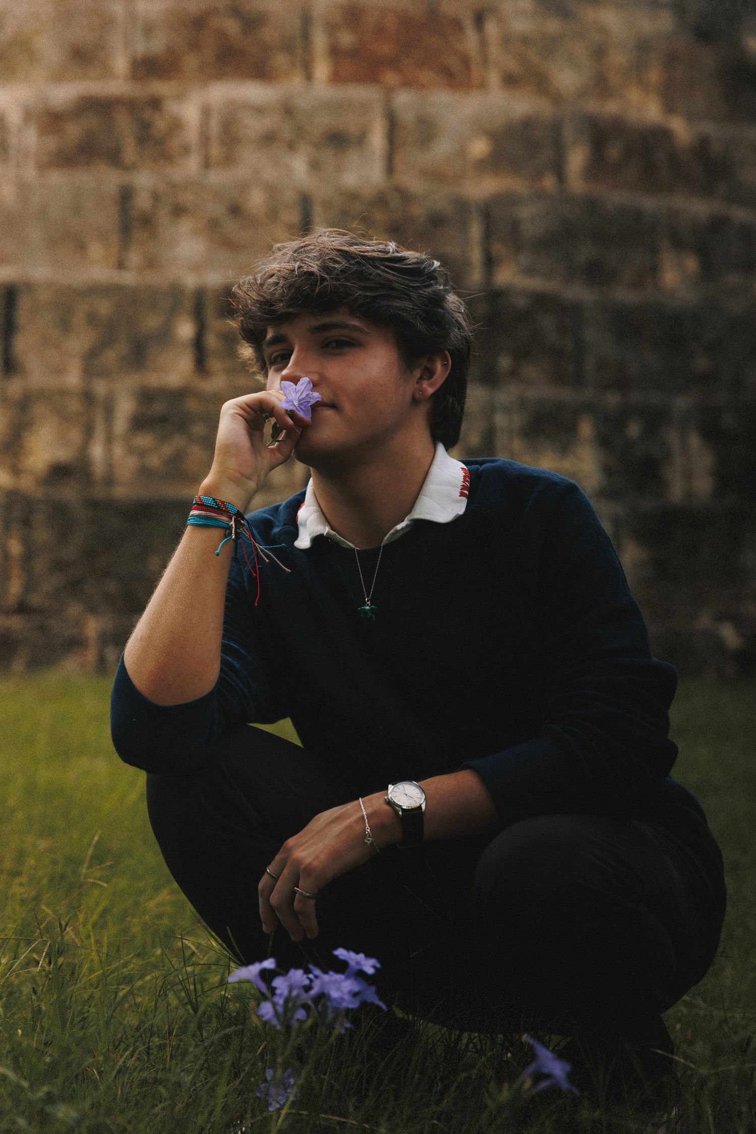 A person in a black sweater crouches near purple flowers against a rustic stone wall at dusk.
