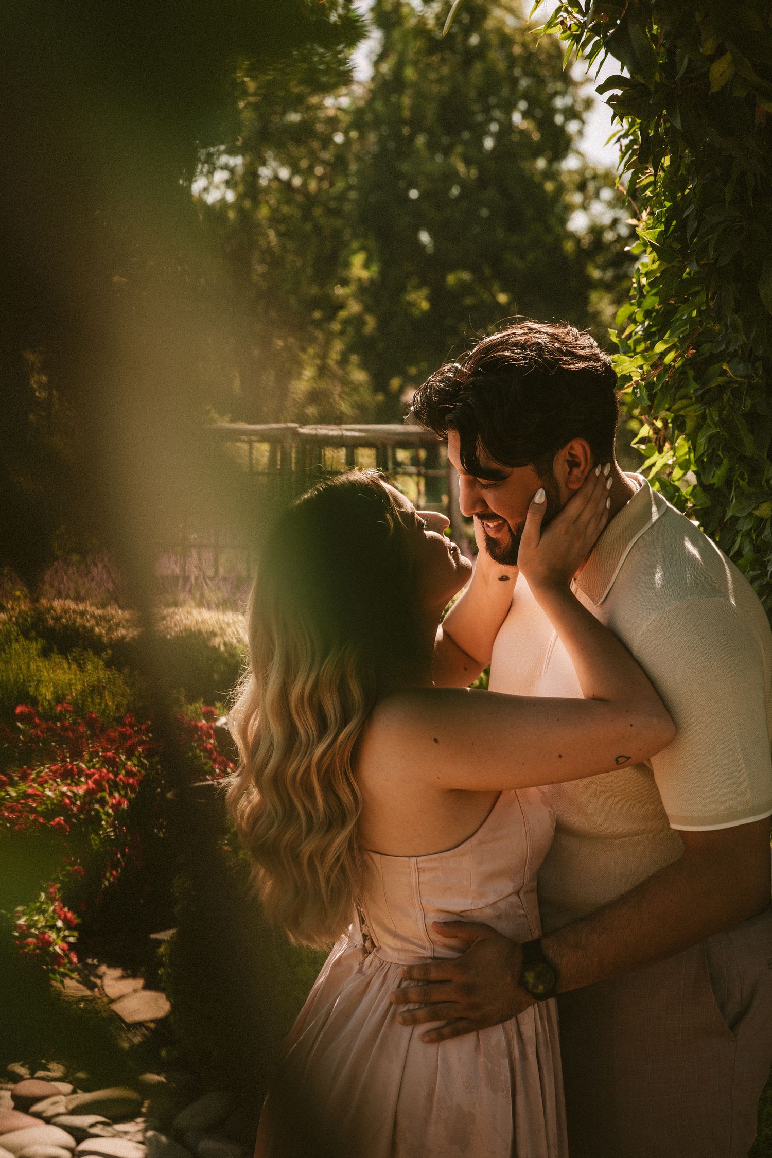 A romantic couple embraces in a sun-drenched garden with soft golden light filtering through trees and flowers.