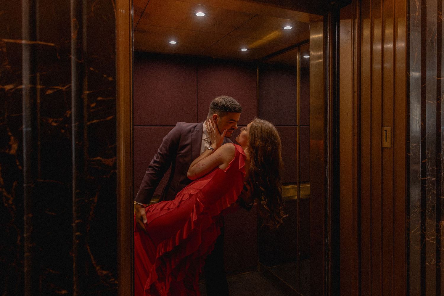 Couple sharing a romantic kiss in an elegant elevator, woman in flowing red dress, moody warm lighting.