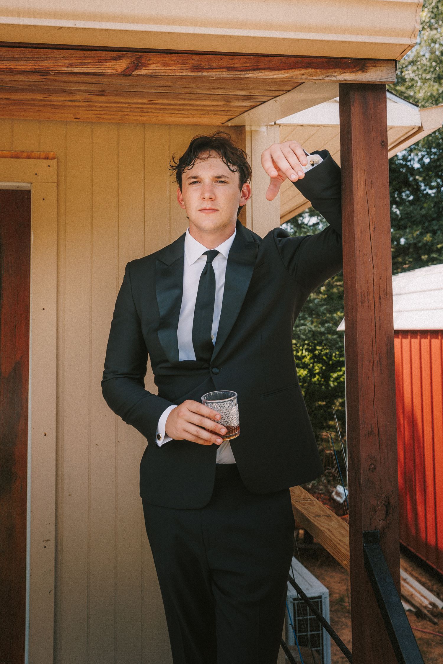 Person in black suit poses casually on wooden porch during formal event.