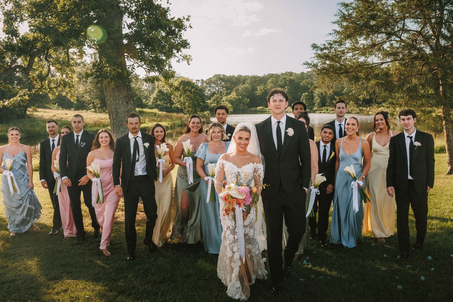 Large wedding party poses for group photos outdoors with a lake in the background.