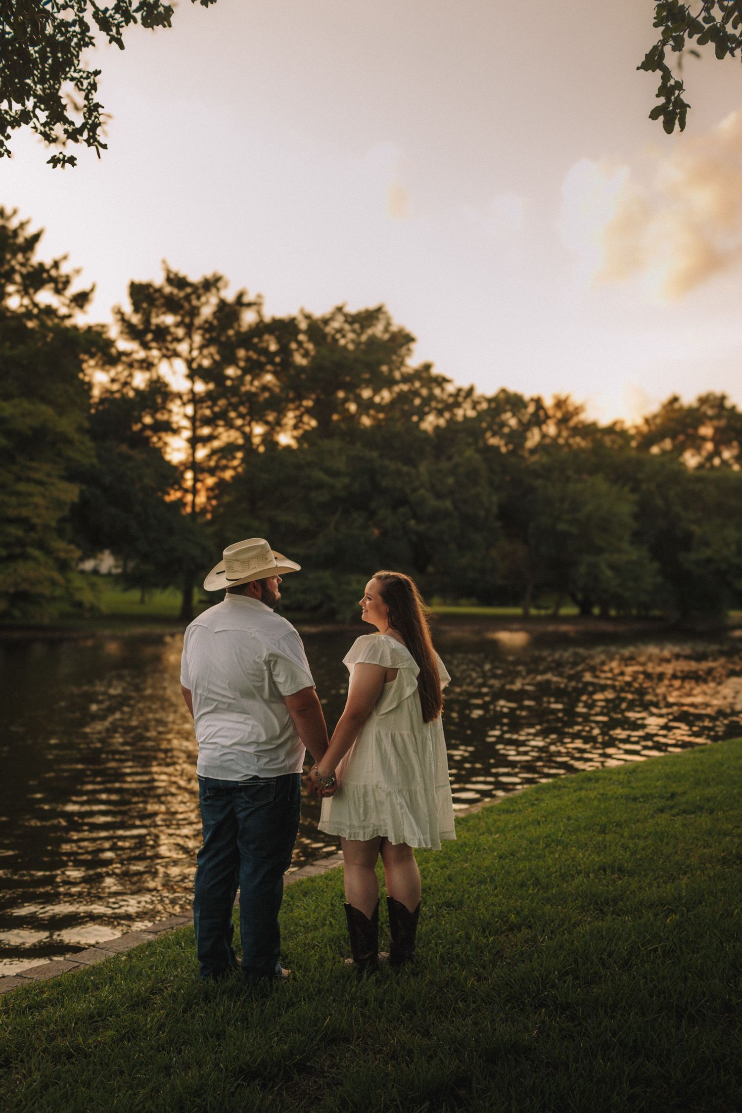 Couple holding hands at sunset by a serene pond with lily pads and trees silhouetted in the golden light.