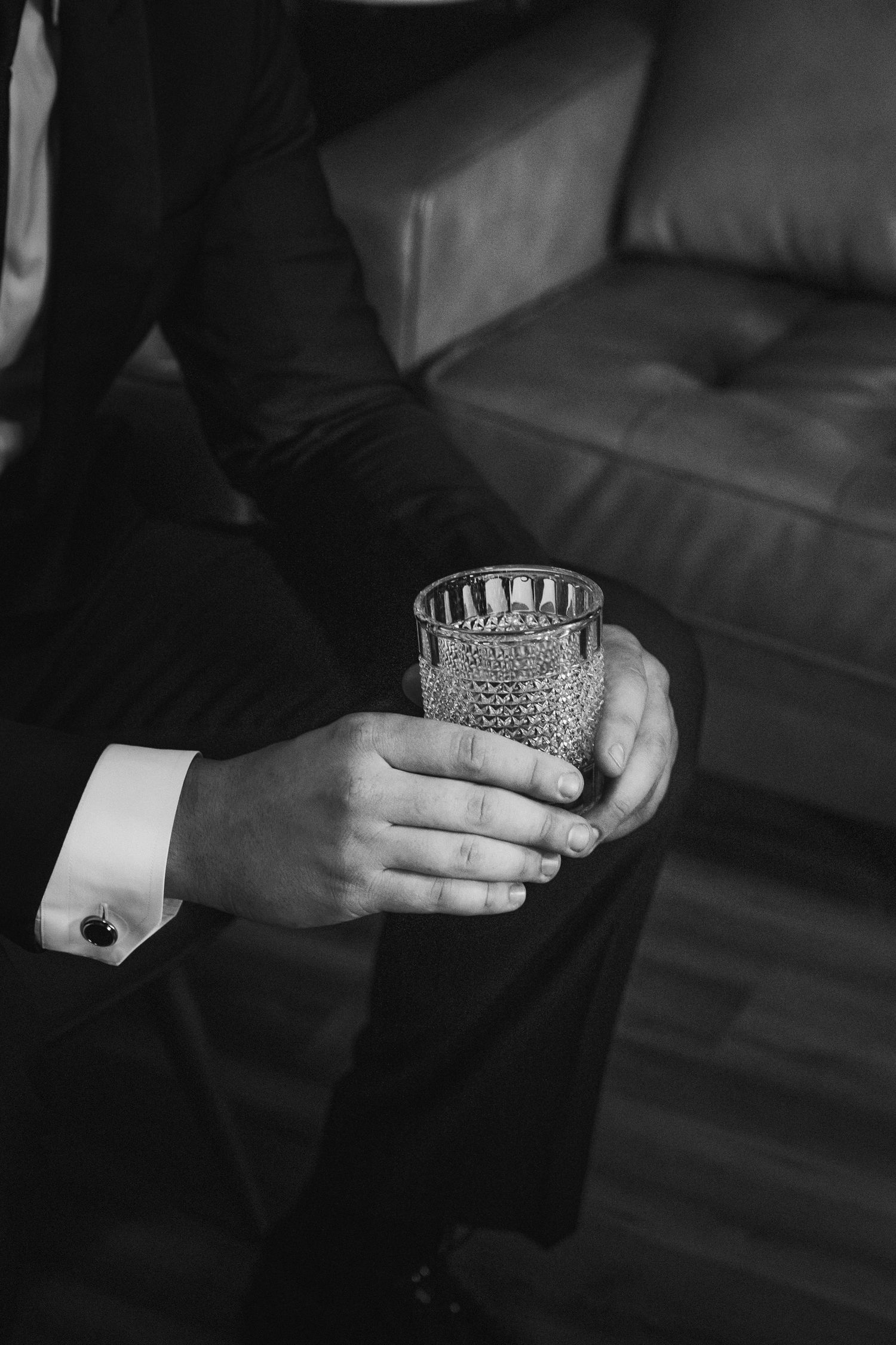 Elegant black and white photo of hands holding crystal whiskey glass with formal attire cuff and cufflink visible.