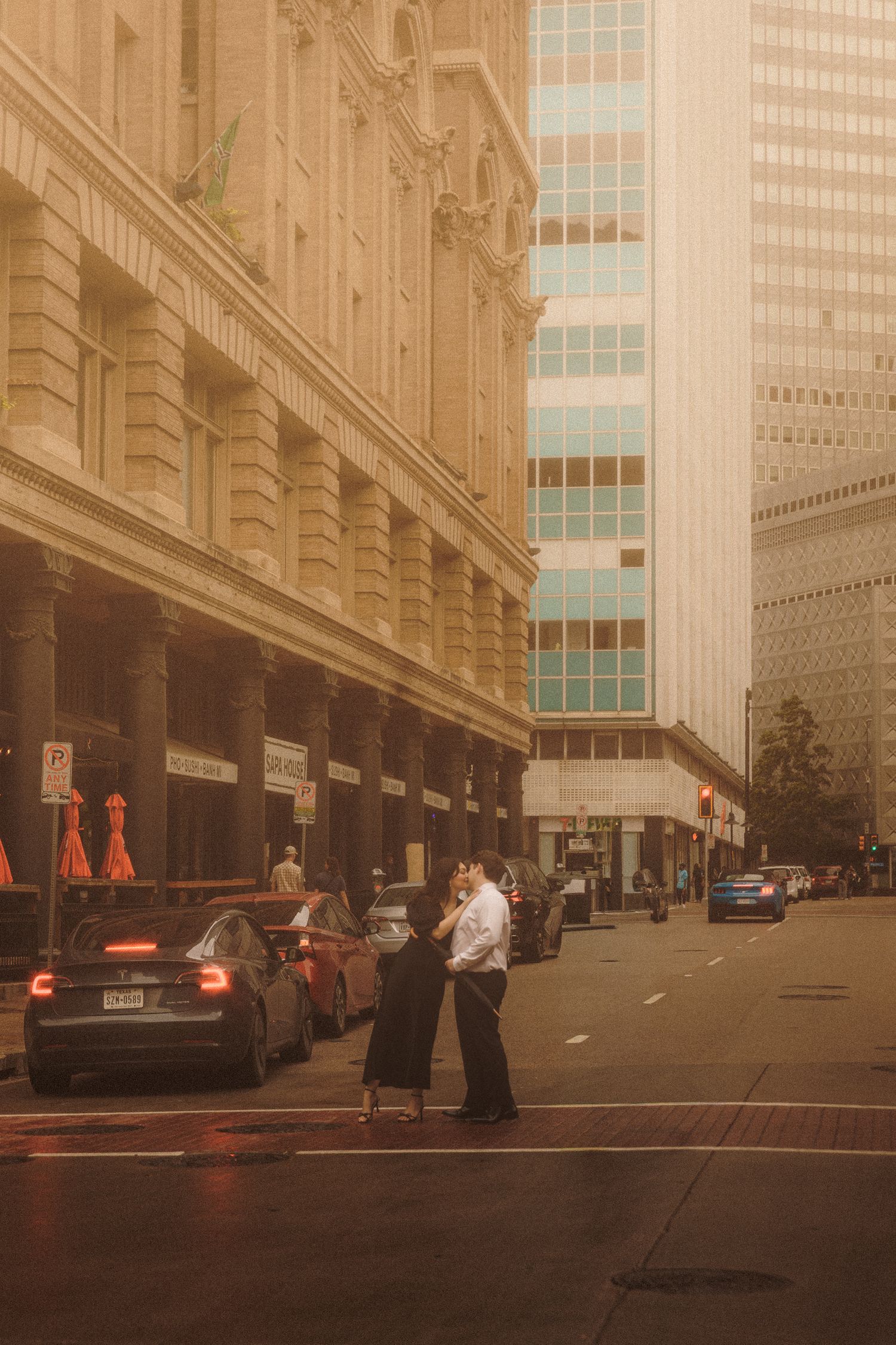 A couple embraces romantically on a city street corner surrounded by classic architecture and parked cars.