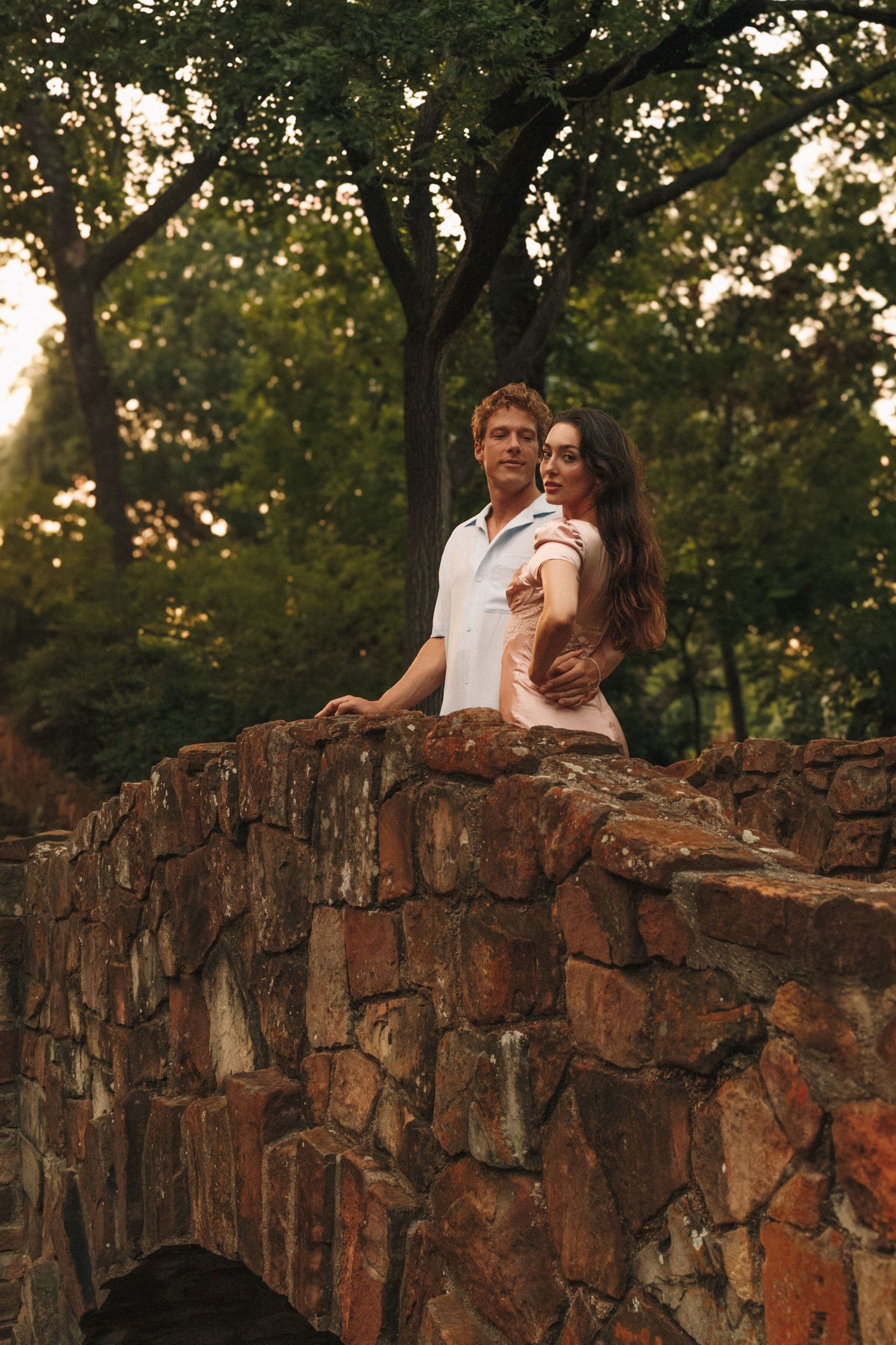 A couple shares an intimate moment while sitting on rustic red rock formations during golden hour in a wooded setting.