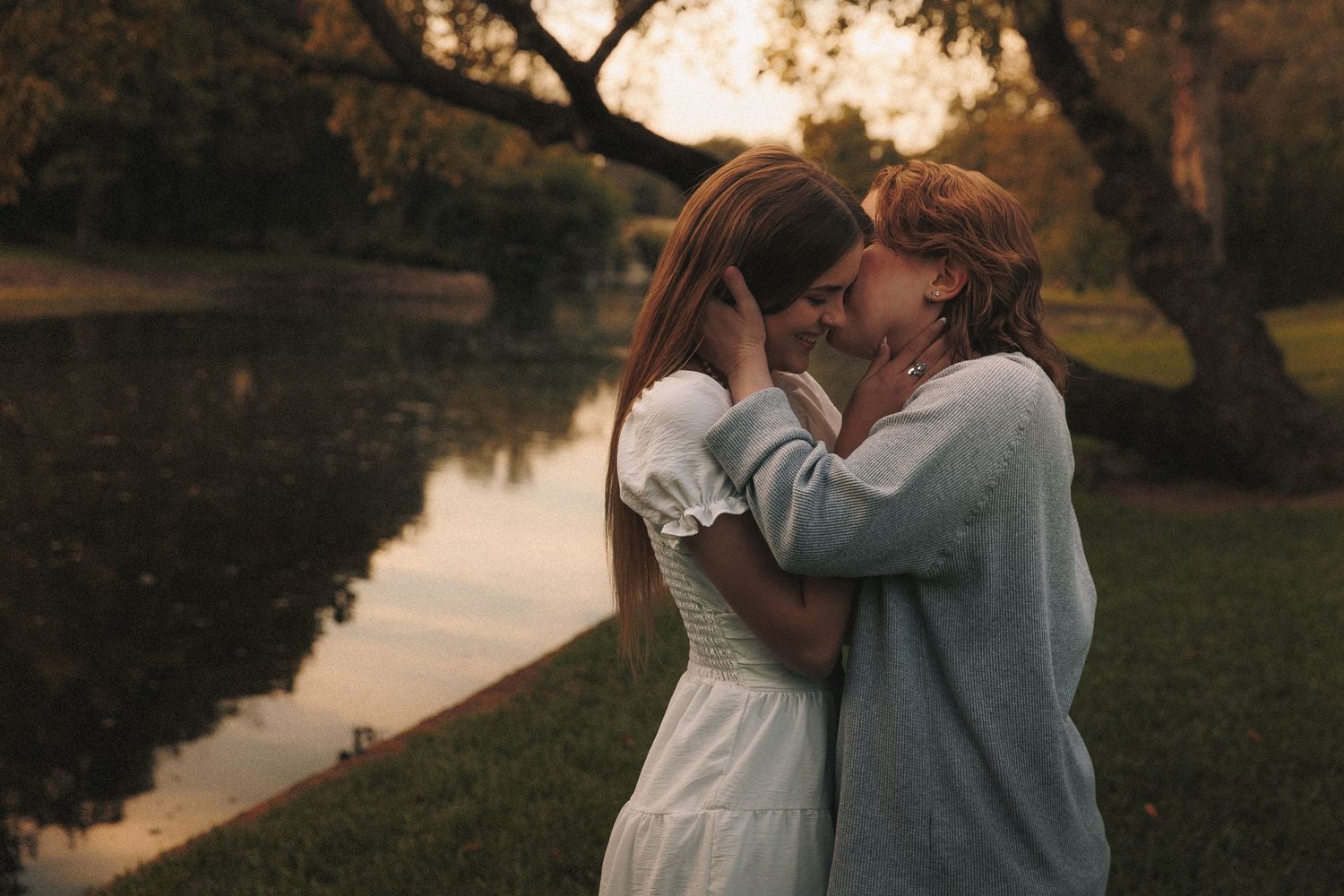 A couple sharing a tender moment by a tranquil lake at sunset with autumn trees in the background.