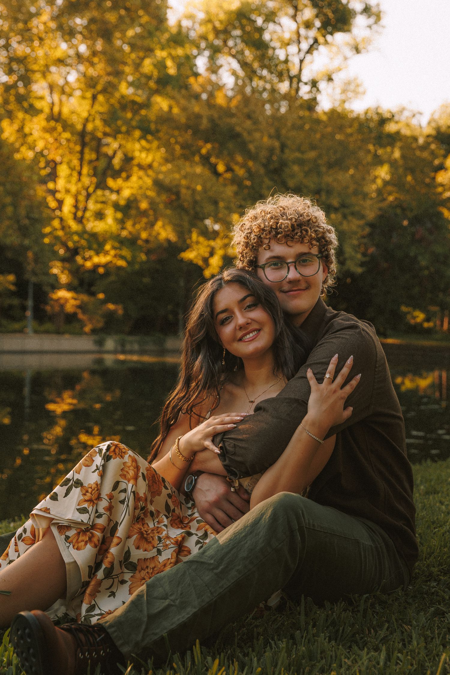 Couple laying together on grass during sunset with autumn foliage in background.