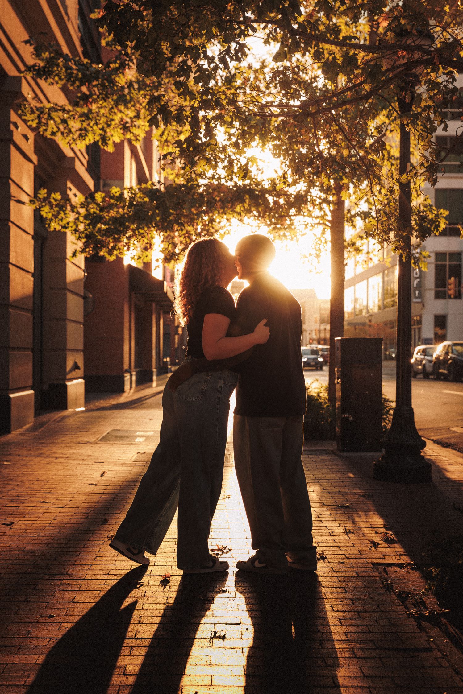 Silhouettes of people against sunset light filtering through trees along a brick-paved walkway.