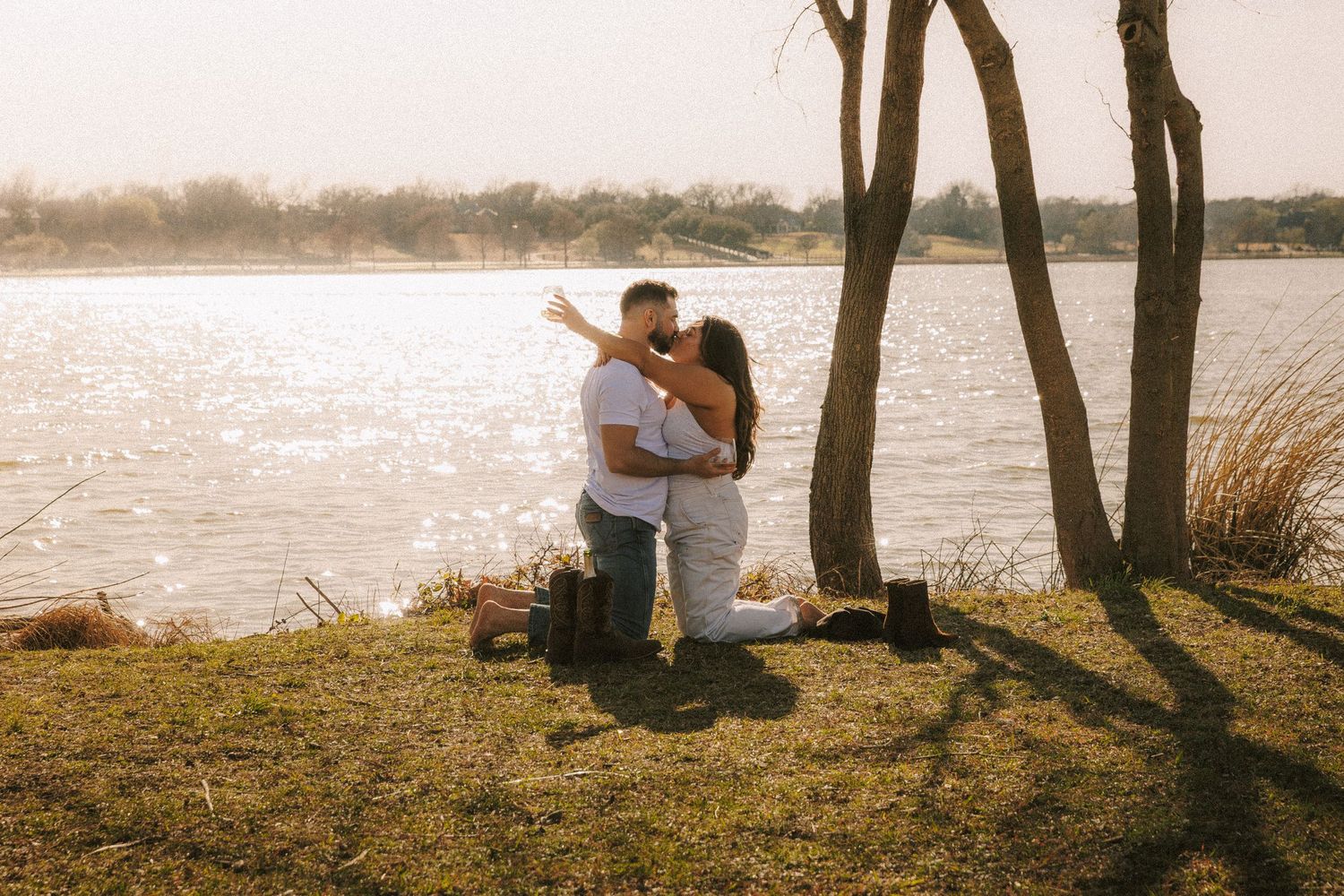 A couple embracing by a lake during golden hour with trees silhouetted against the water.