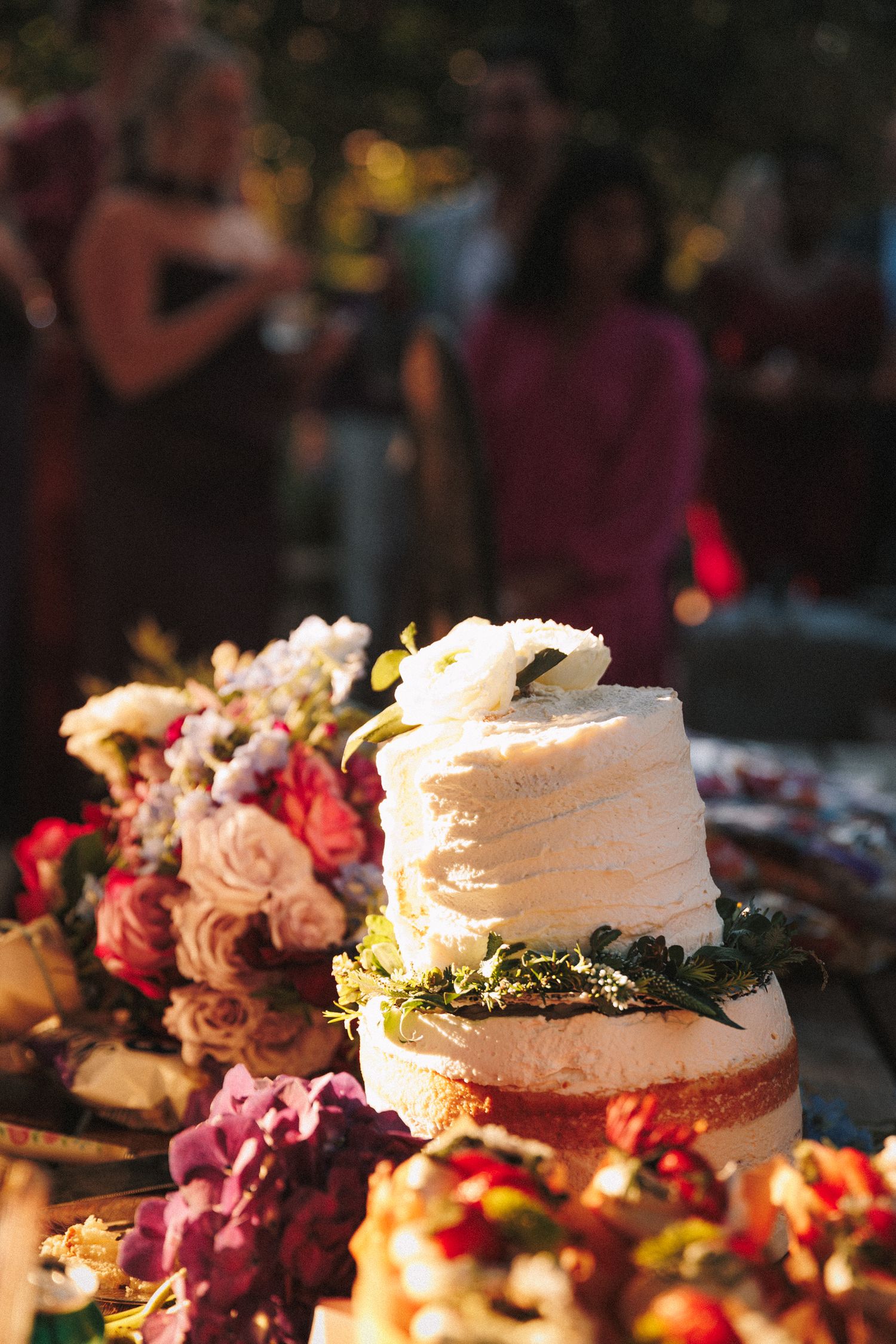 Elegant two-tier white naked wedding cake adorned with greenery and white flowers at reception.