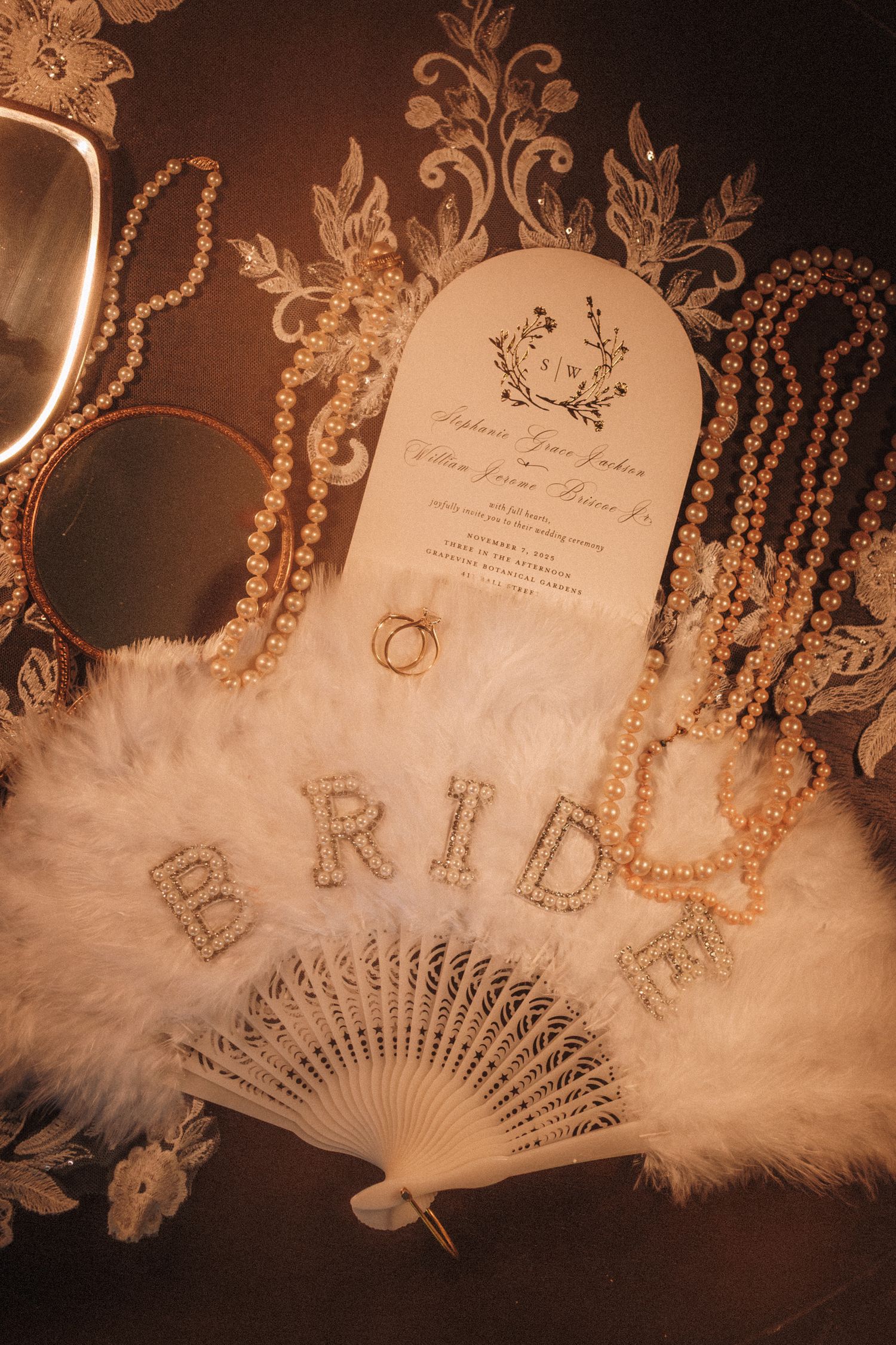 Bridal flat lay with feather fan spelling BRIDE, pearl necklaces, wedding invitation, and gold ring.