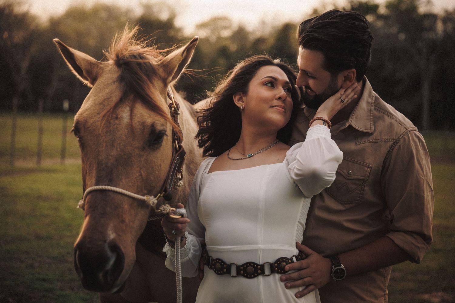 Couple embracing near a horse on a farm during a glowing golden hour sunset.