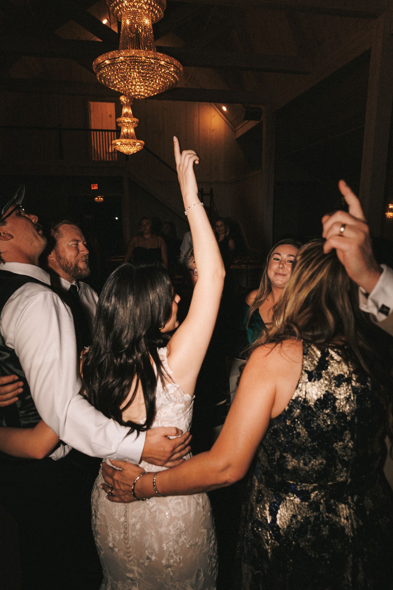 Bride dancing with guests under chandelier at lively indoor wedding reception celebration.