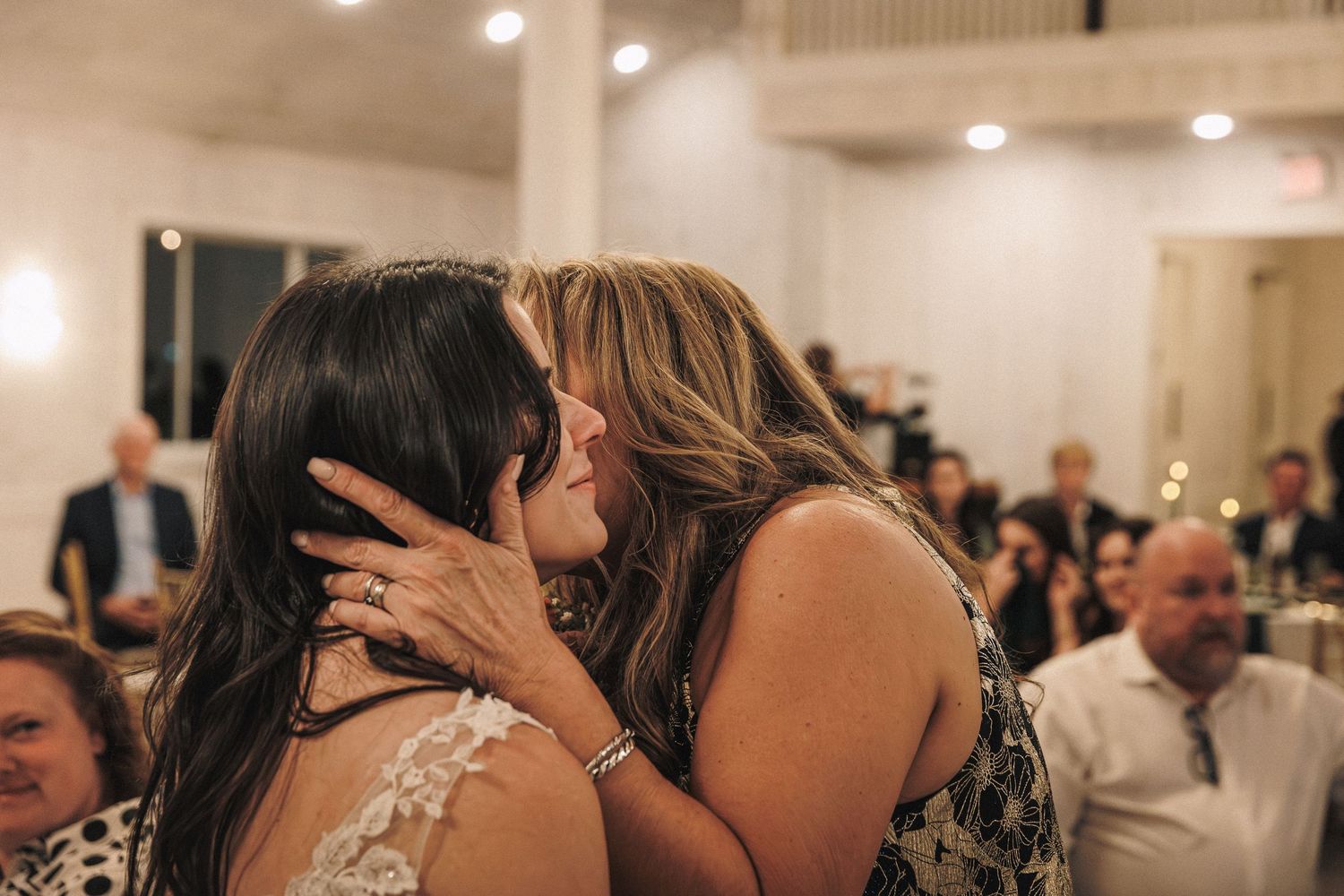 Emotional mother and bride sharing heartfelt kiss at wedding reception celebration.
