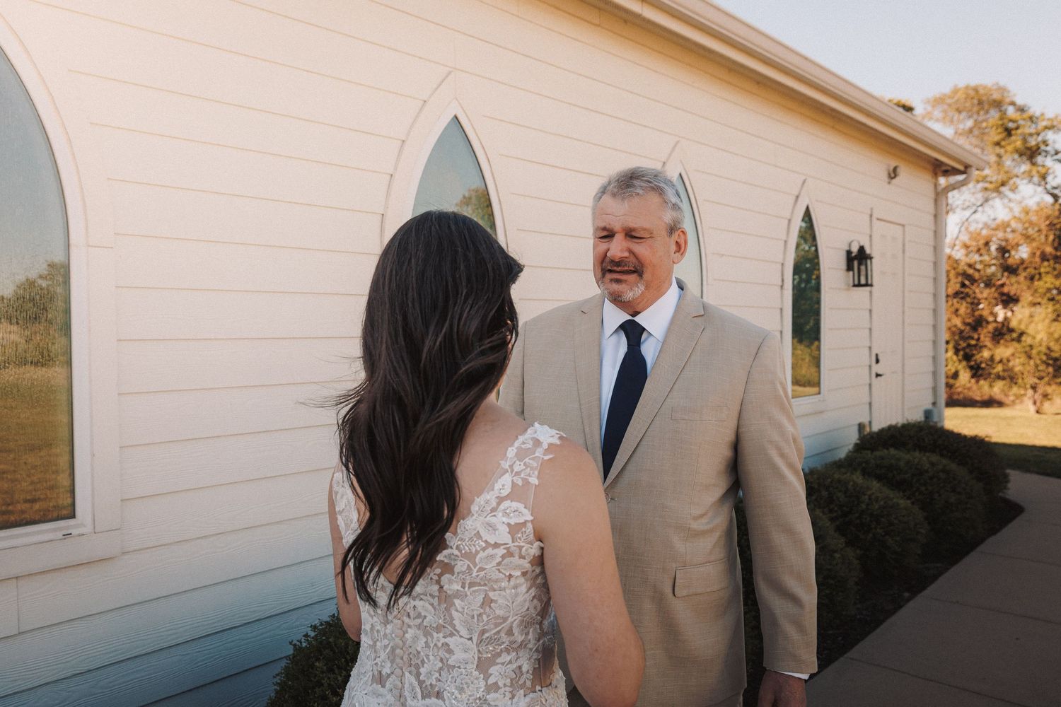 Emotional father smiling at bride in lace wedding dress outside white chapel.