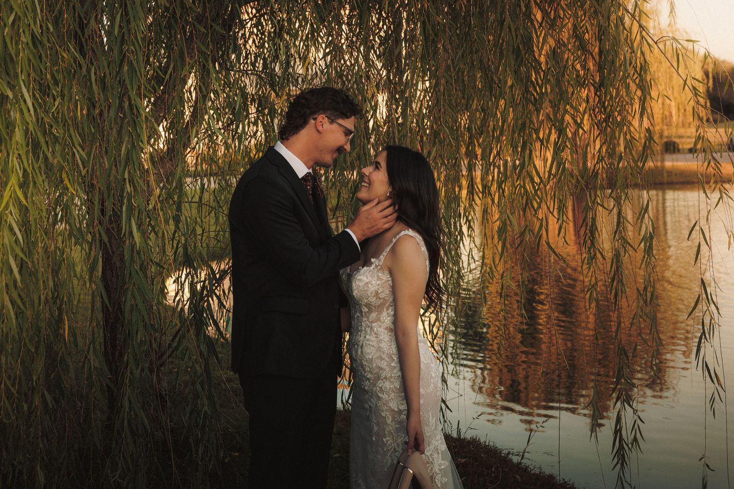 Bride and groom share a romantic moment under weeping willow tree by a serene lake.