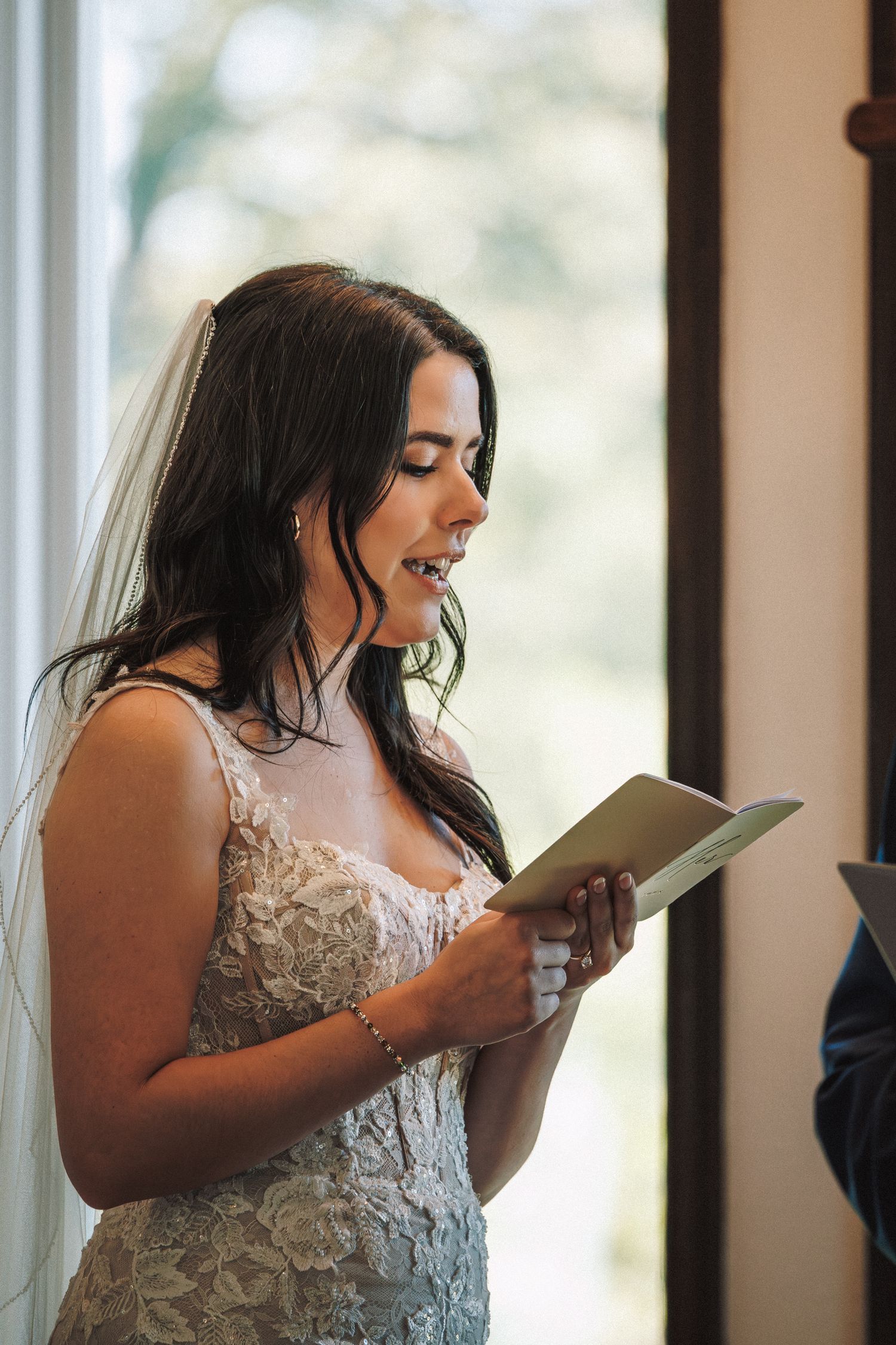 Bride in lace gown and veil reads heartfelt wedding vows from a card during the ceremony.
