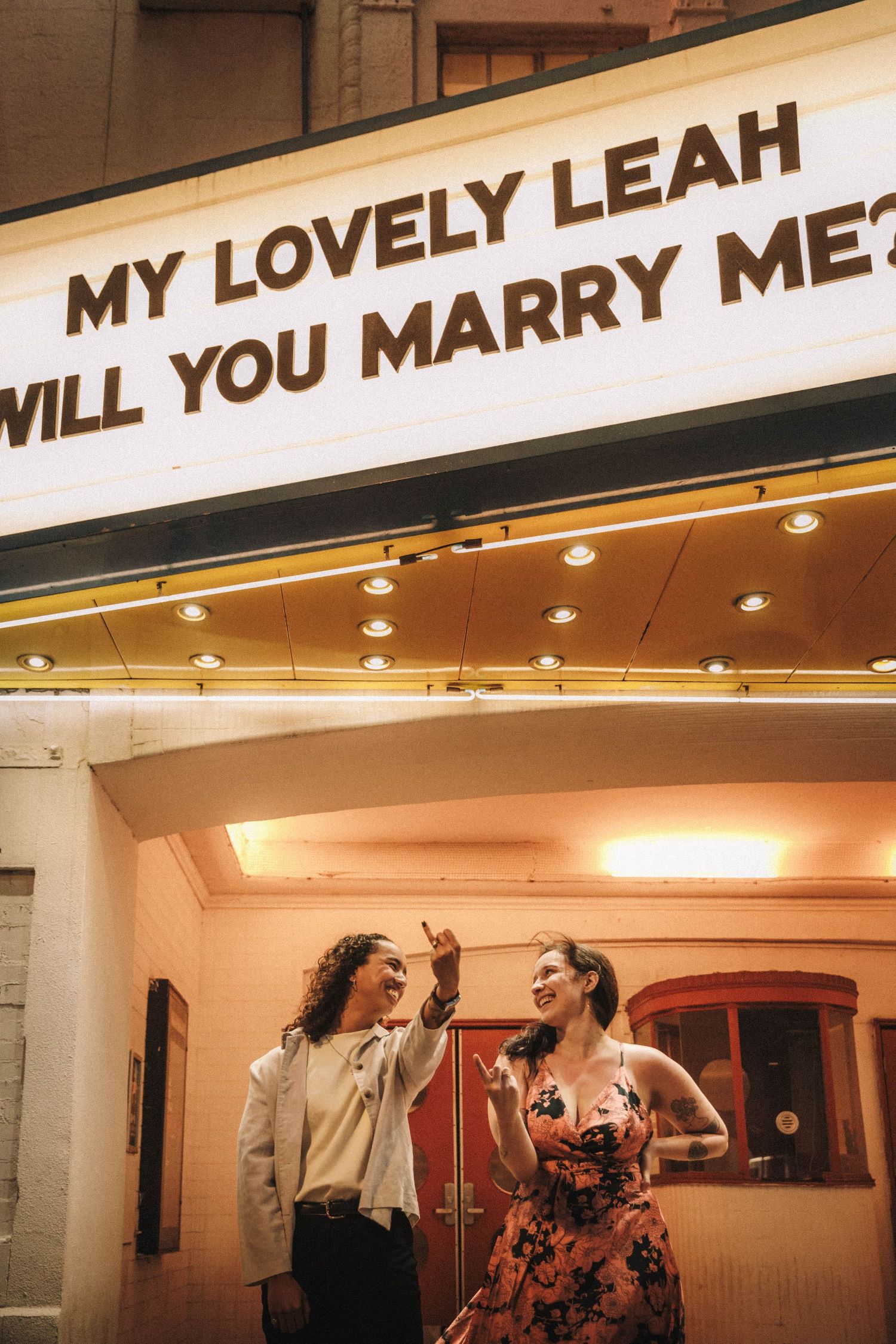 Joyful couple laughs together under glowing marriage proposal theater marquee sign.