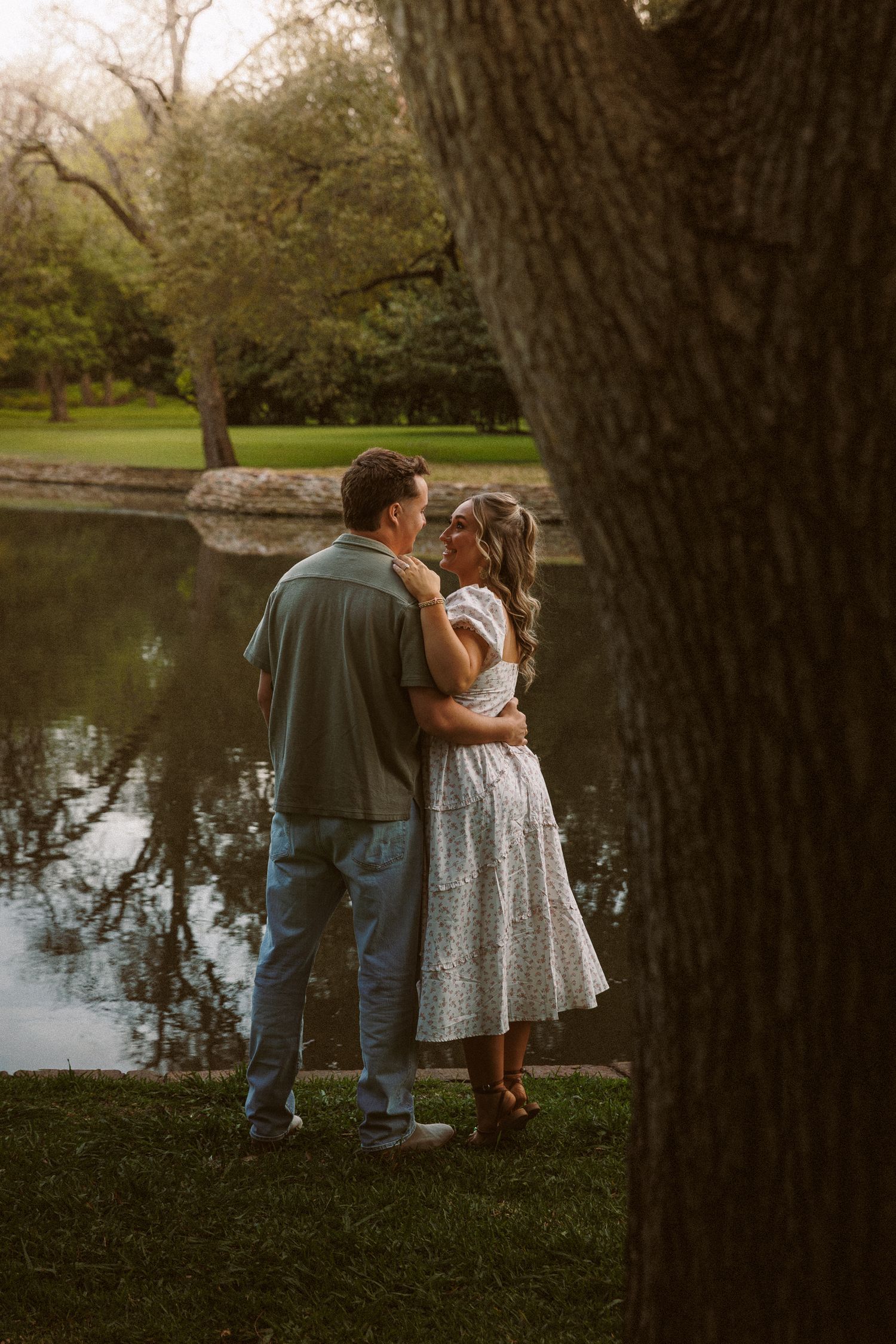 Couple embracing near a serene reflective pond surrounded by trees in soft evening light.