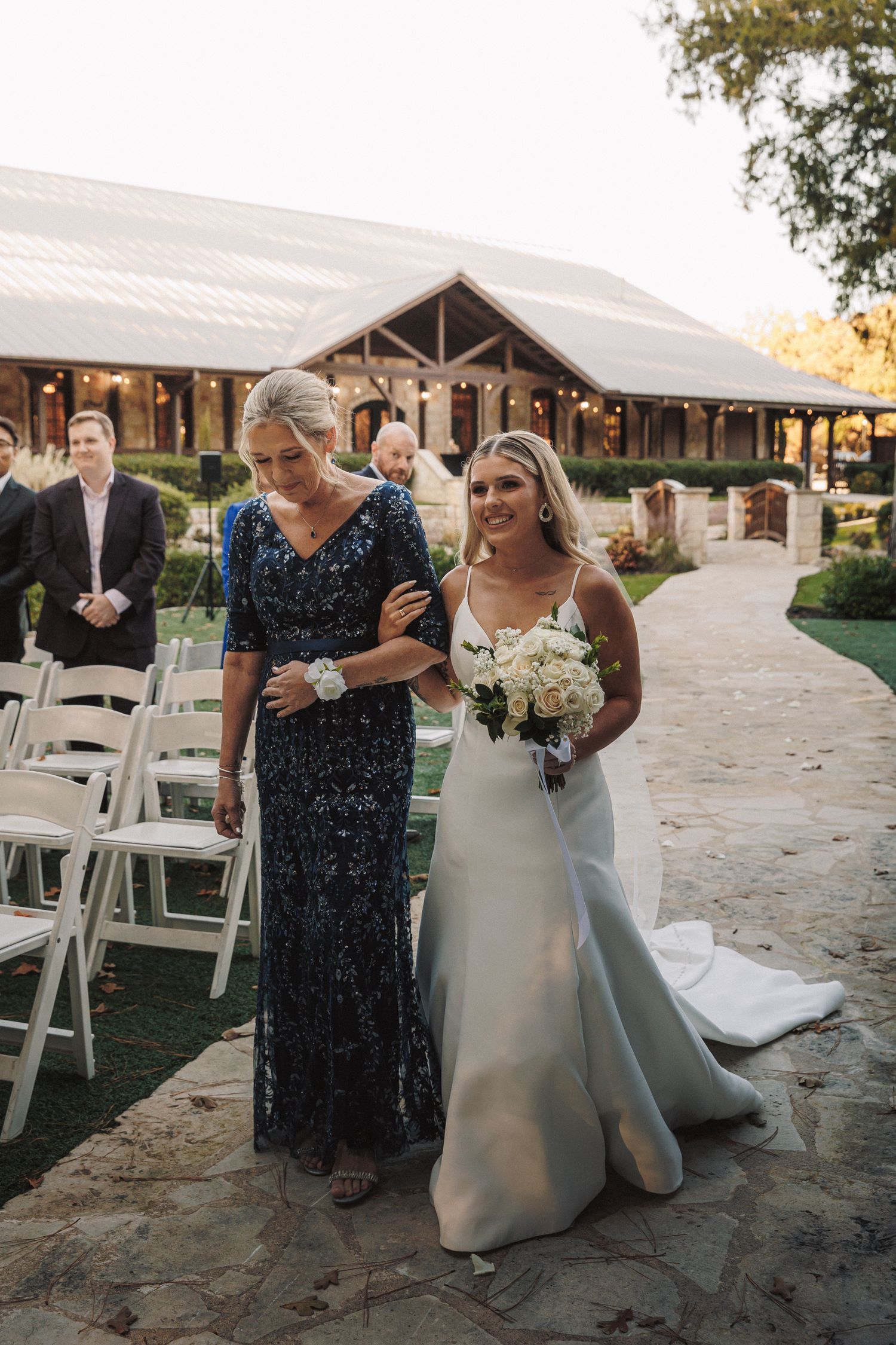 Bride in white gown holding bouquet walks down aisle with escort at outdoor wedding.