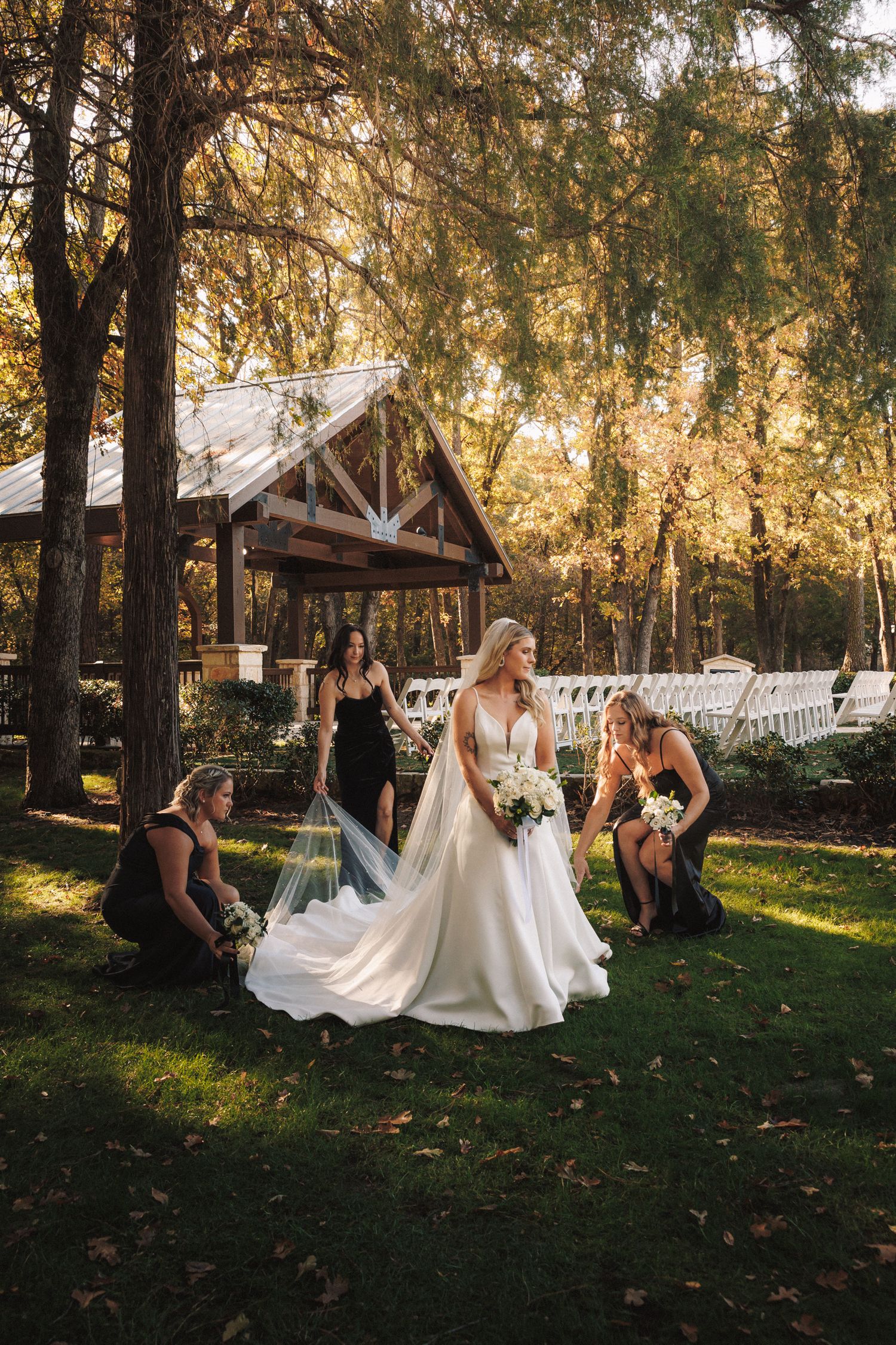 Bridesmaids in black dresses arranging the train of a bride's white gown in an autumn garden.
