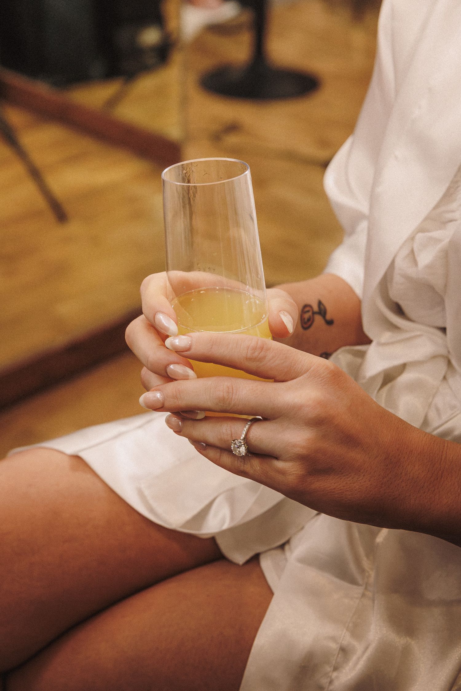 Bride in white robe holding champagne flute with engagement ring visible on her hand during wedding preparations.