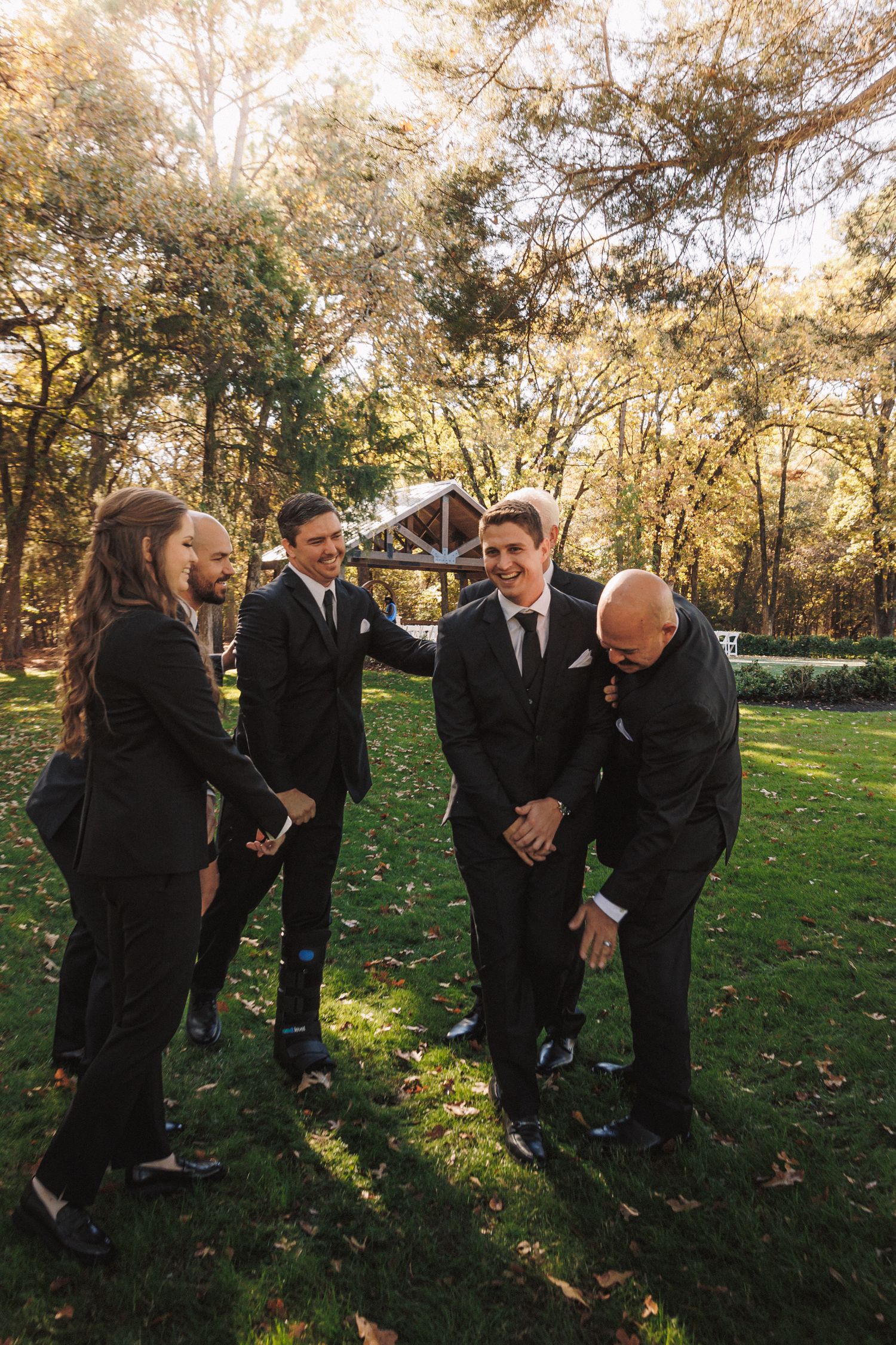 Groomsmen in black suits laughing together outdoors in a sunlit garden during a wedding celebration.