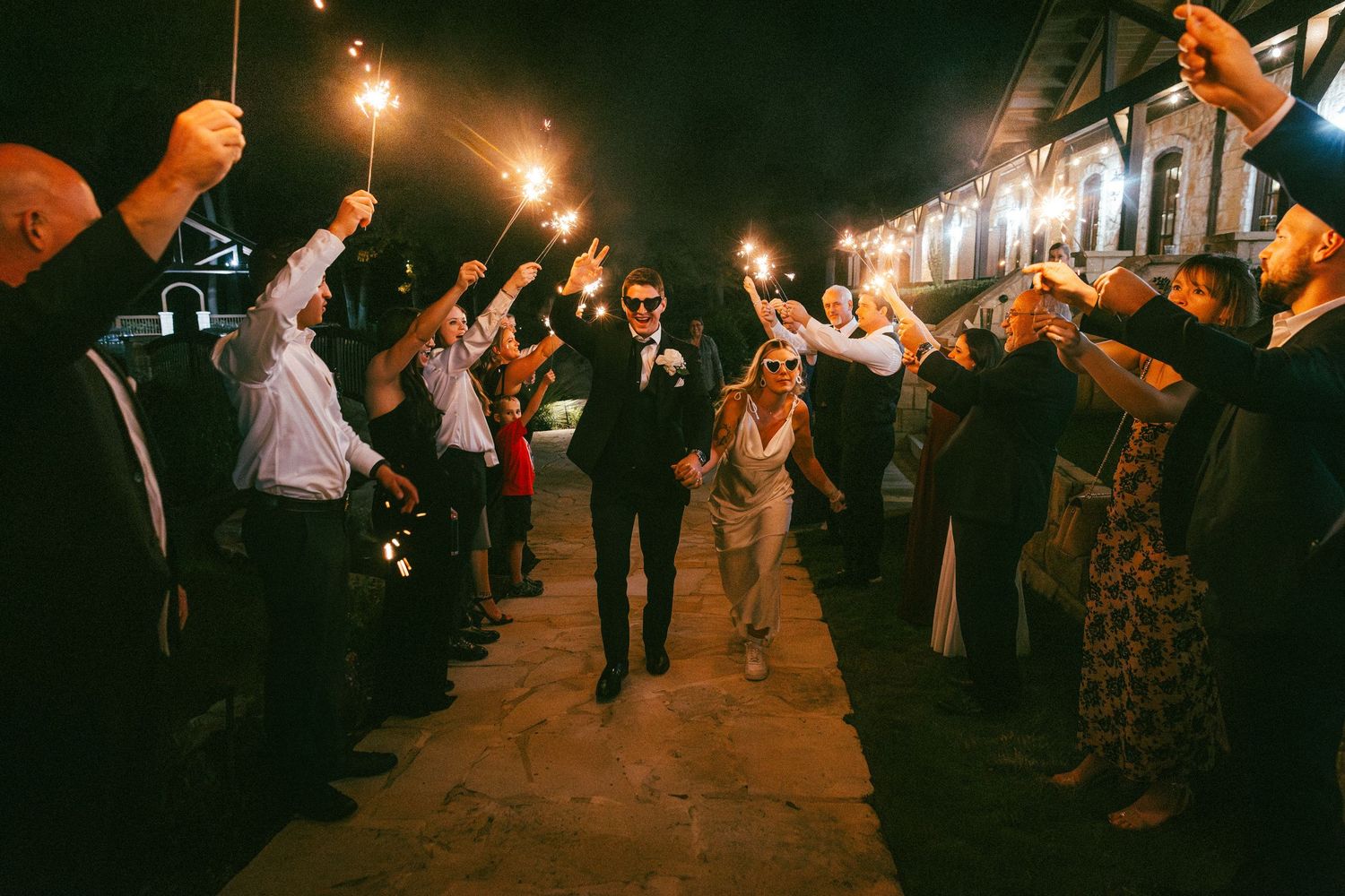 Newlyweds exit through sparkler tunnel surrounded by cheering guests at night wedding sendoff.